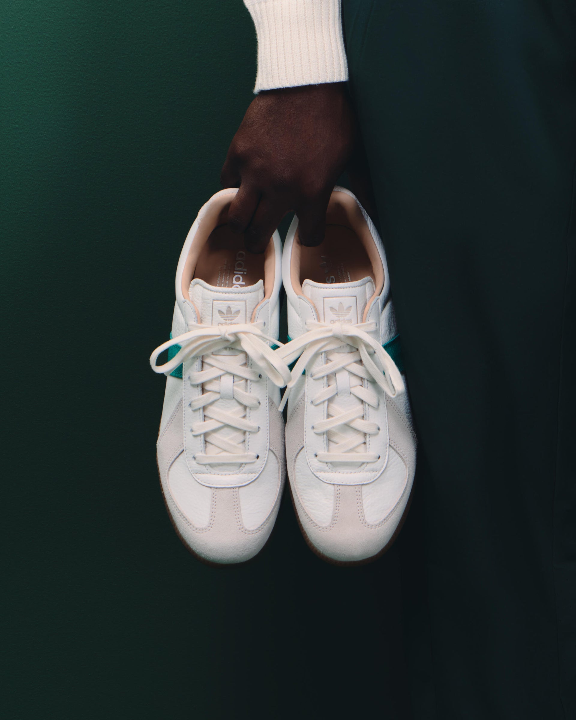 Hand holding a pair of white leather adidas BW Army sneakers with a green side stripe against a dark green backdrop