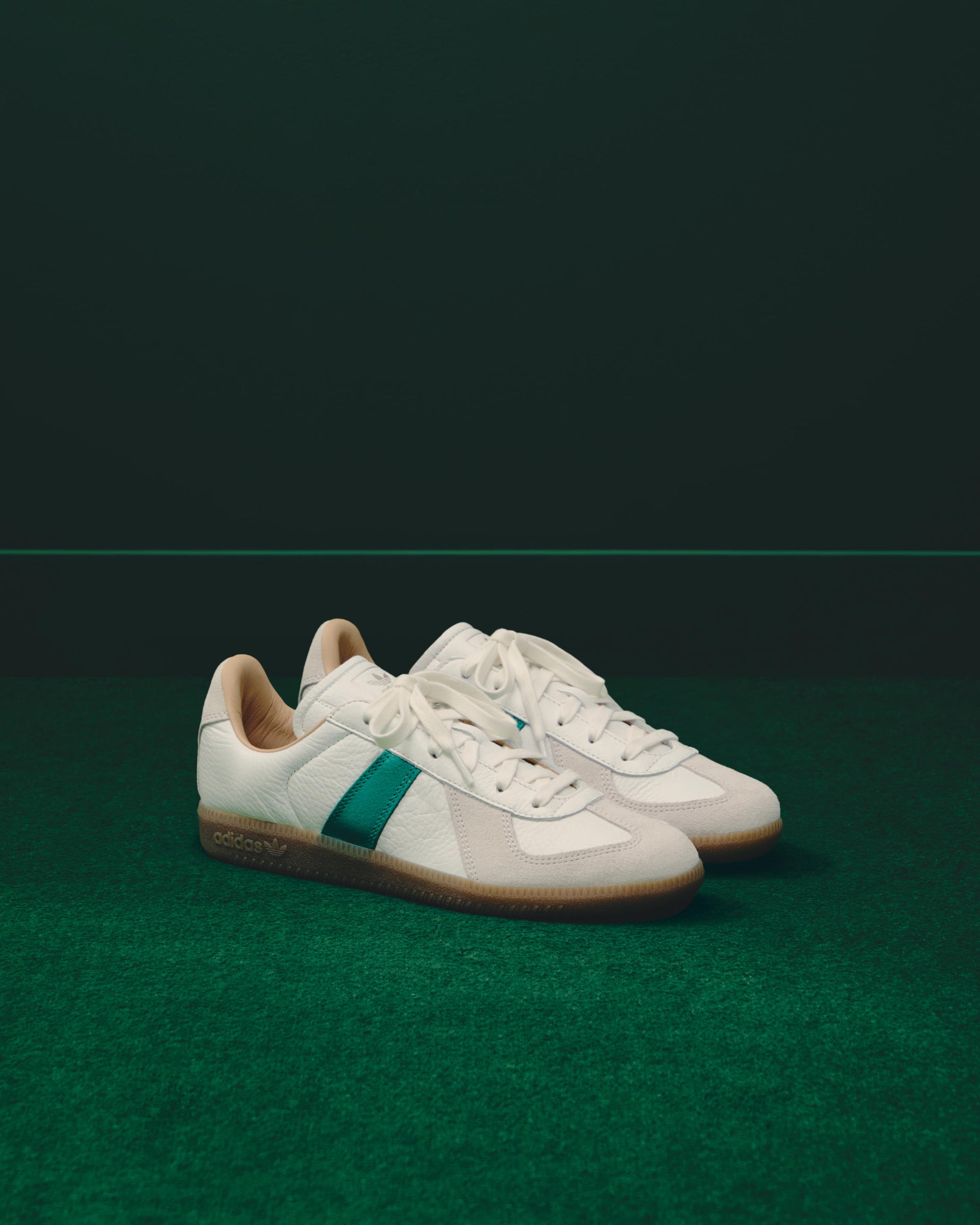White leather adidas BW Army sneakers with a green stripe and gum sole displayed on green turf against a dark green backdrop