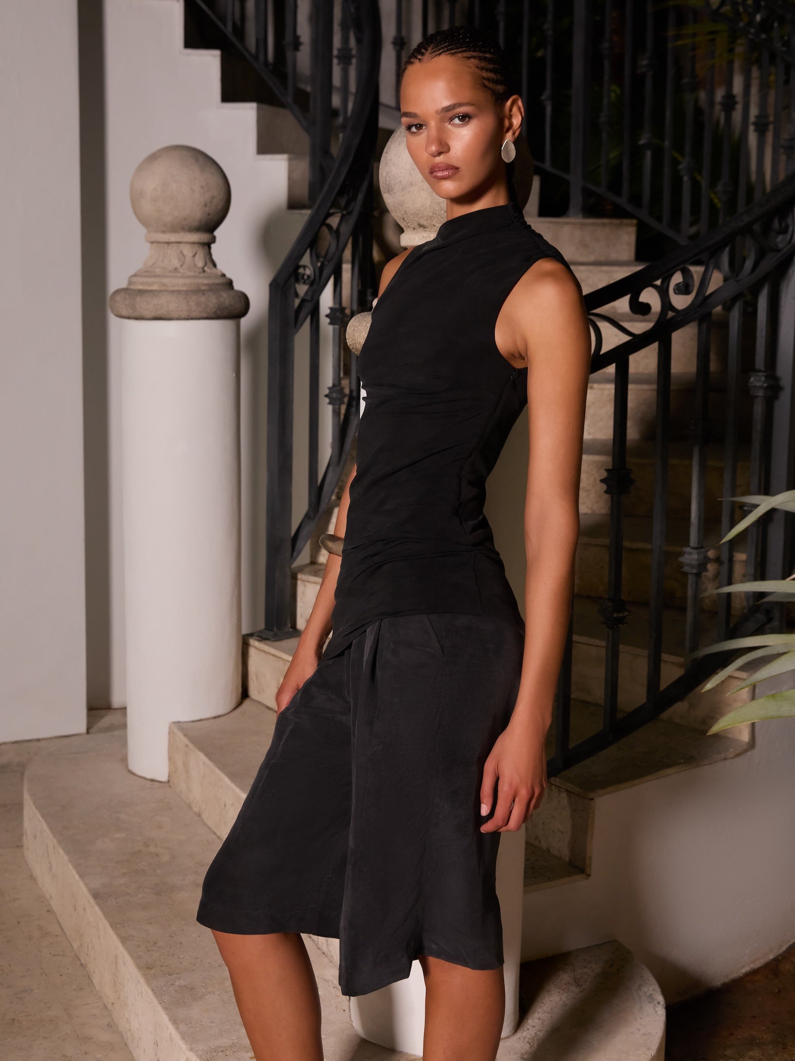 Woman in a black sleeveless mock-neck top and knee-length shorts, side view leaning on an ornate staircase railing