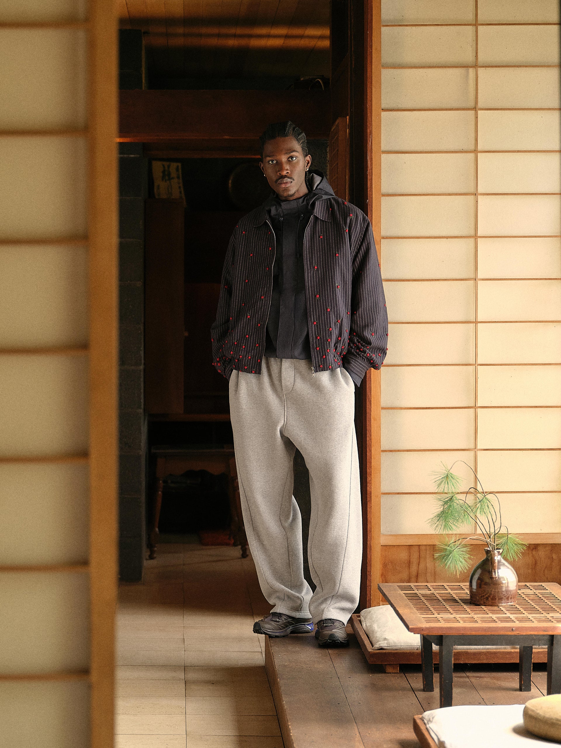 Charcoal pinstripe rose-embroidered jacket and grey sweatpants on model standing in doorway, full-length