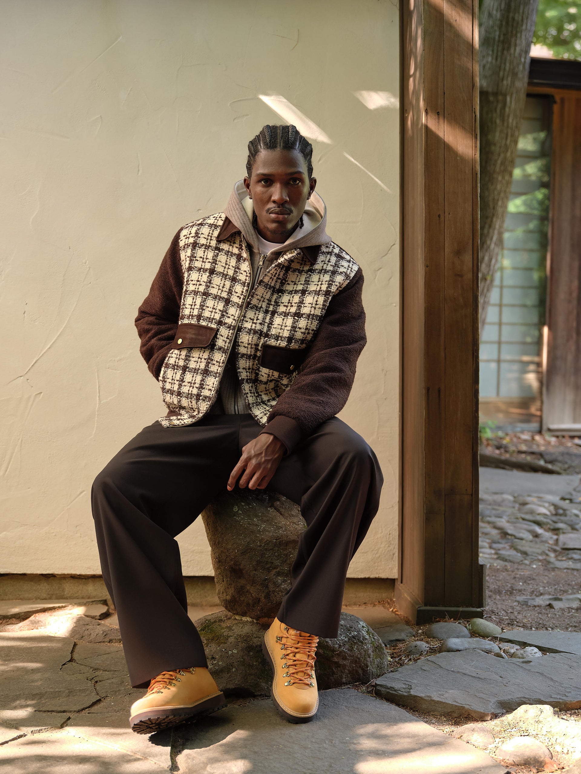 Model seated on a rock in a cream-and-brown checked tweed jacket, brown trousers and tan lace-up hiking boots outside a stucco house