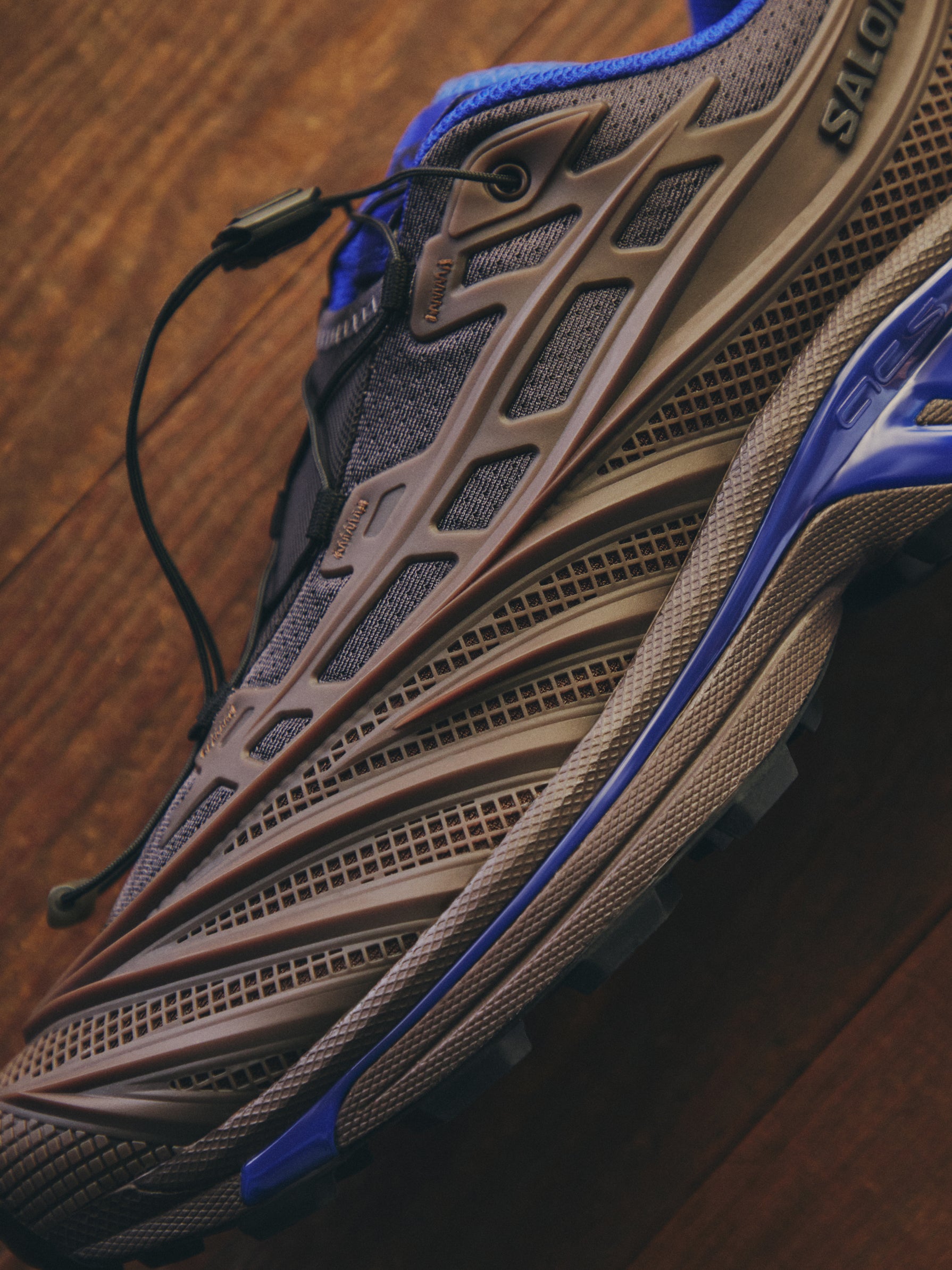 Close-up detail of a taupe and blue Salomon trail sneaker showing the sculpted midsole and mesh upper on a wood floor