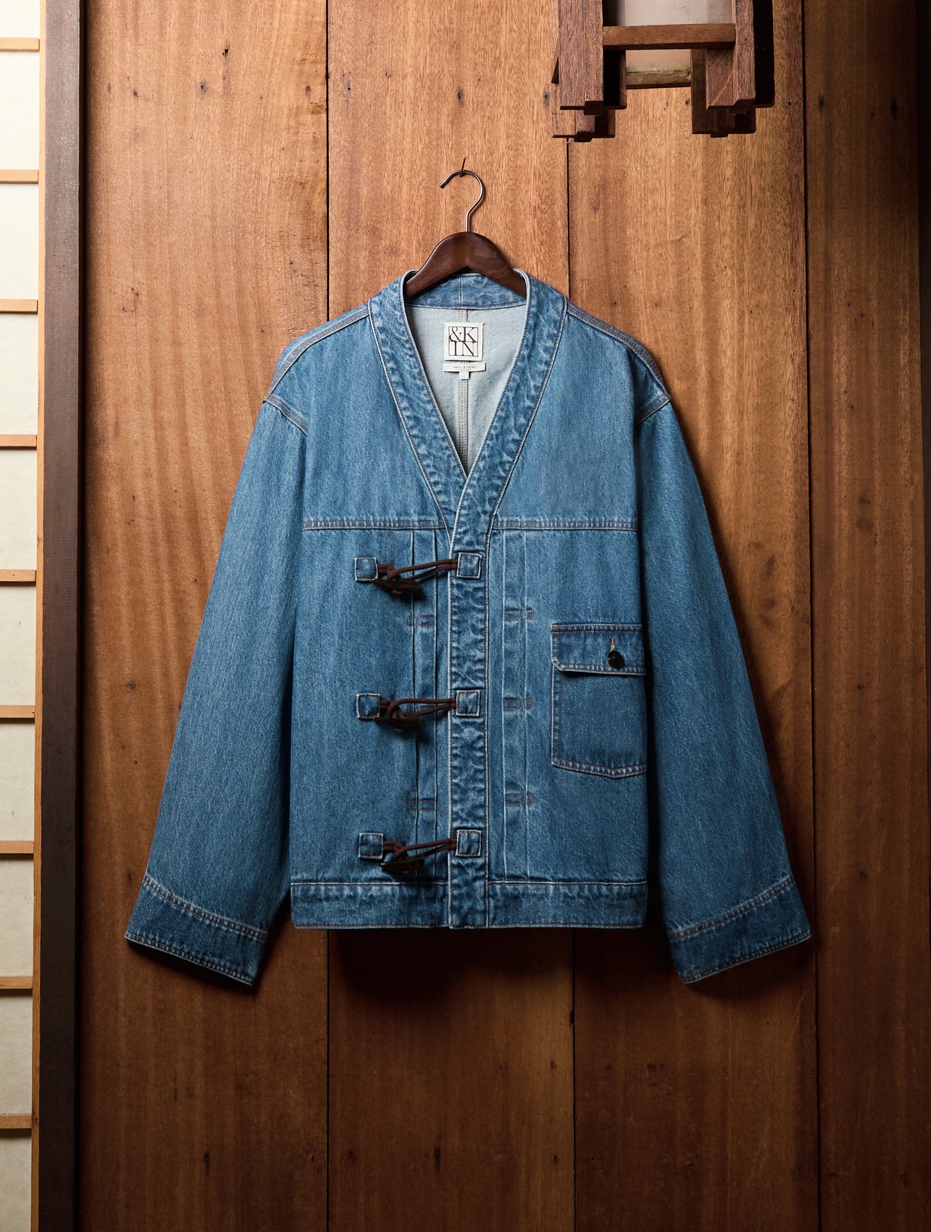 Washed-blue denim kimono jacket with leather toggle closures and a patch pocket on a wooden hanger against a wood-panel wall