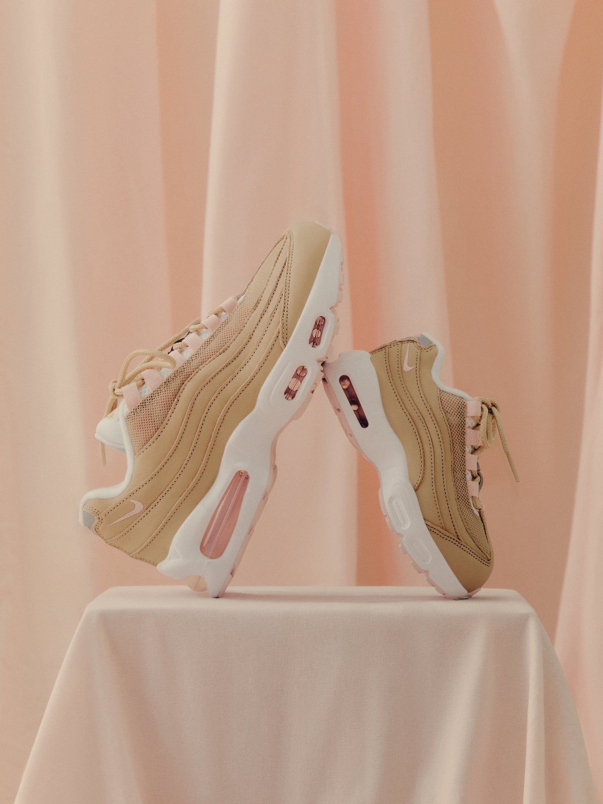 Pair of Nike Air Max 95 in tan and pink displayed on draped blush-pink fabric at Kith