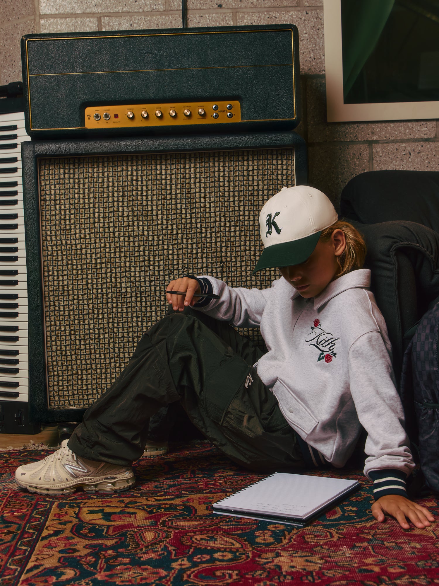 Blond boy on a Persian rug against green amps in a white-and-green Kith cap and heather gray Kith rose-graphic hoodie