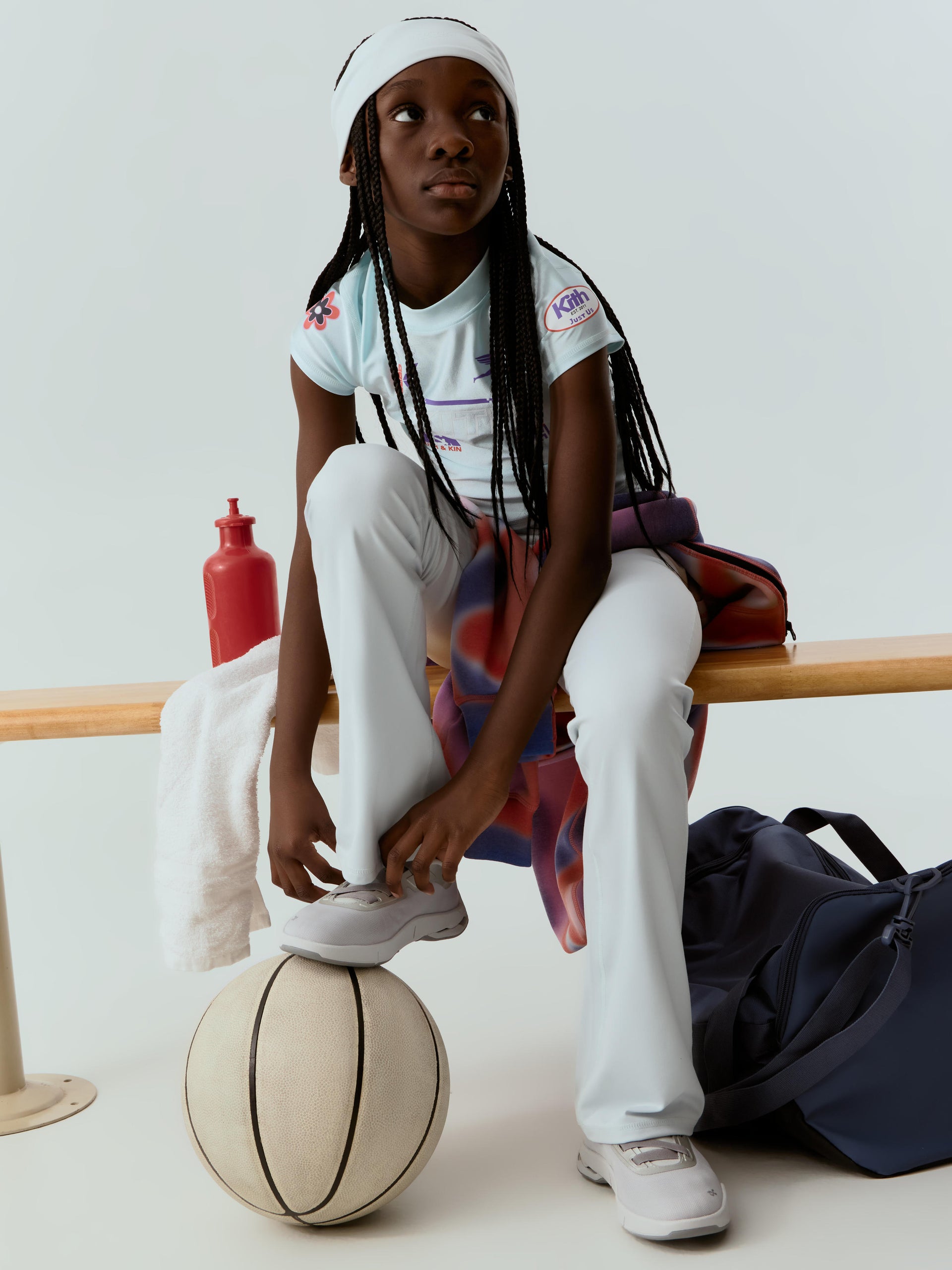 Young Black girl basketball player on a wood bench in a mint Kith graphic tee, foot resting on a basketball, navy duffel bag alongside