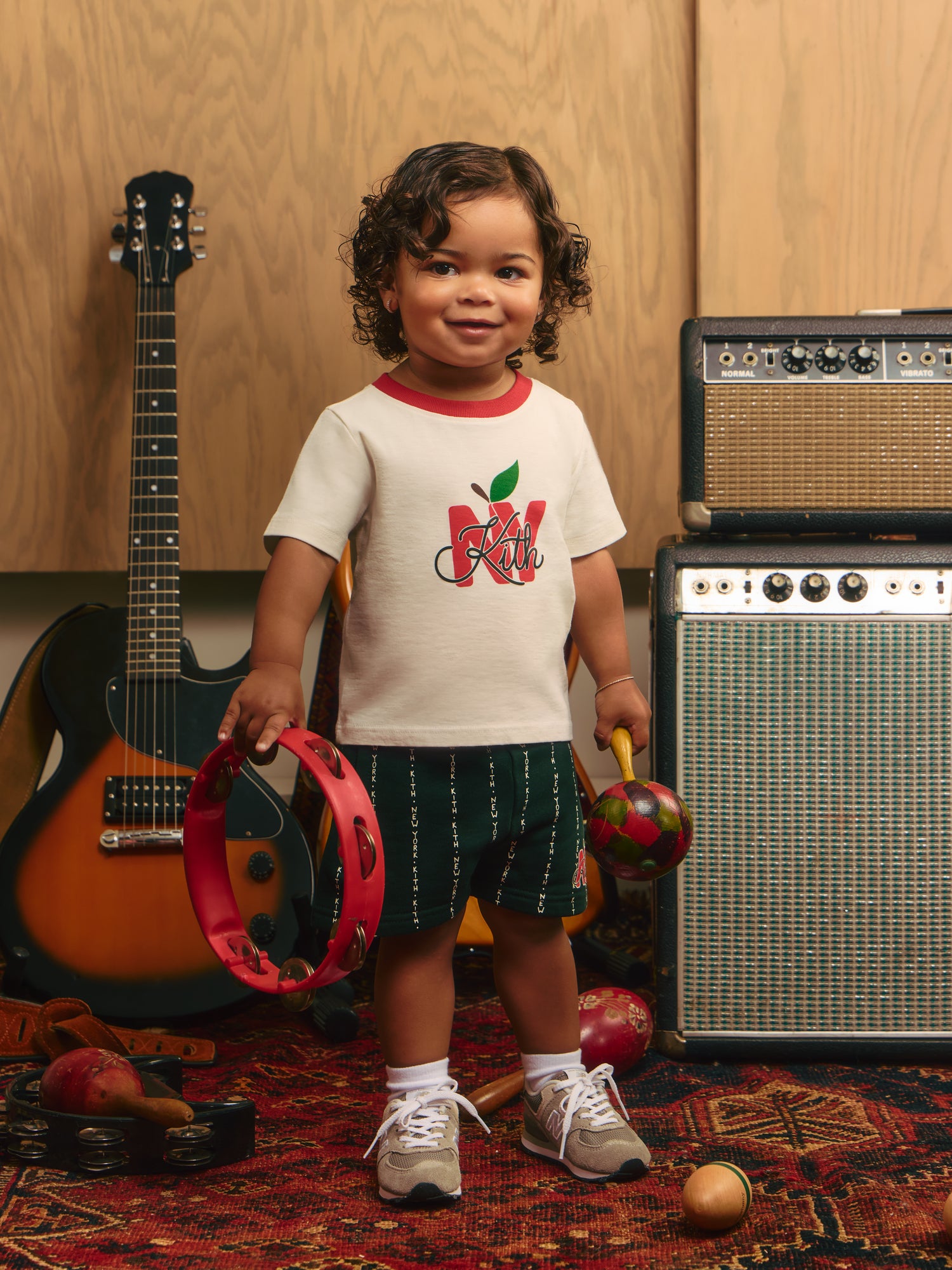 Toddler in a cream Kith NY apple ringer tee and green Kith New York shorts holding a red tambourine and maraca by a guitar and amps