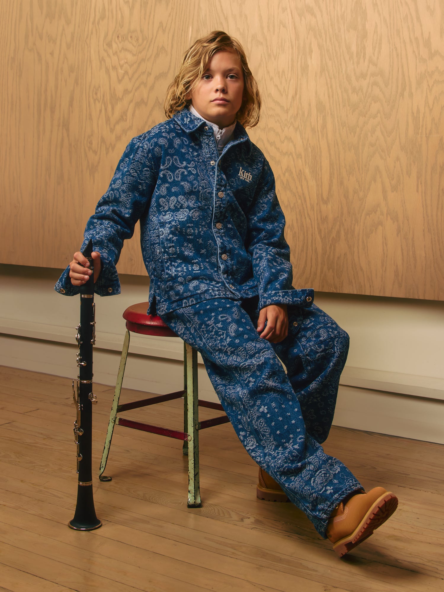 Blond boy on a stool in an indigo Kith New York paisley denim jacket and jeans with Timberland boots, holding a clarinet