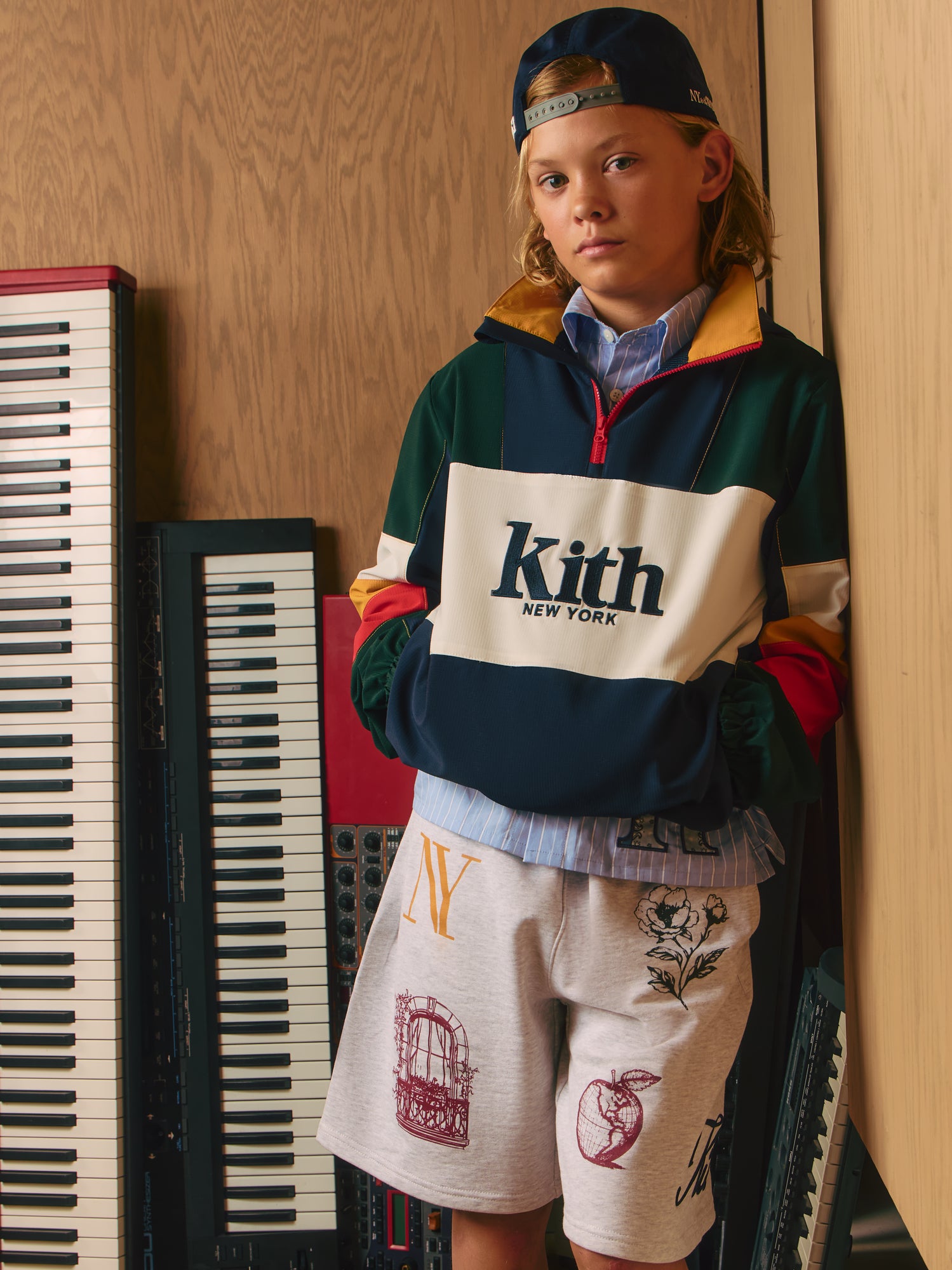 Blond boy in a backwards cap and green-navy-red colorblock Kith New York quarter-zip with gray Kith graphic shorts by stacked keyboards