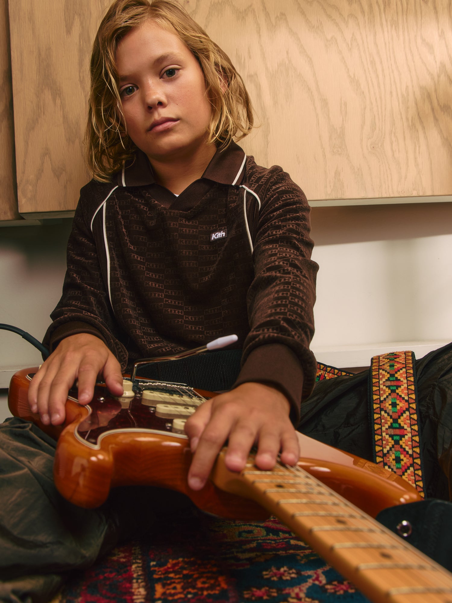 Blond boy holding a wood-finish electric guitar in a brown Kith monogram velour polo with white piping on a Persian rug
