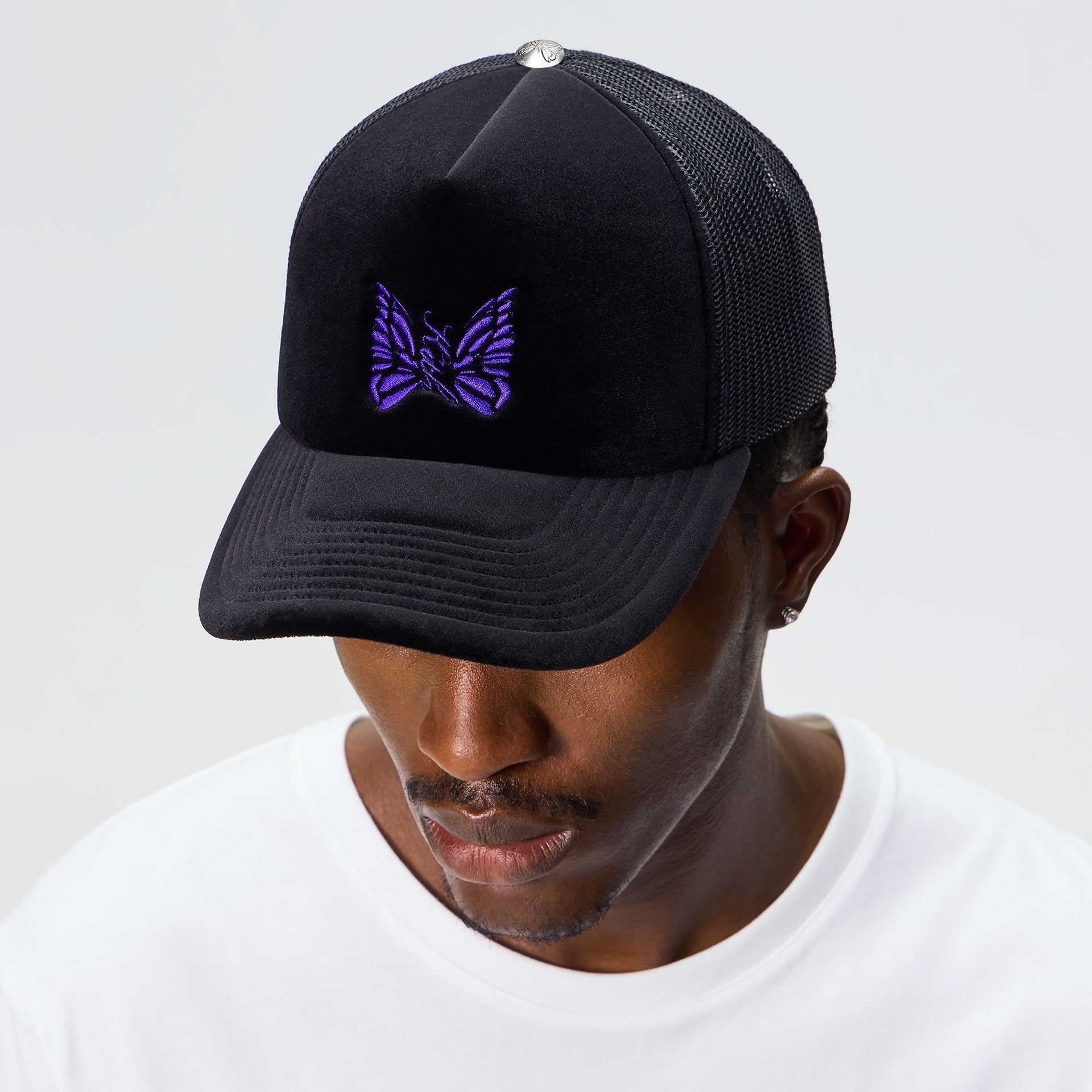 Man wearing the Kith for Needles Velour Nolan trucker cap in black with purple butterfly logo