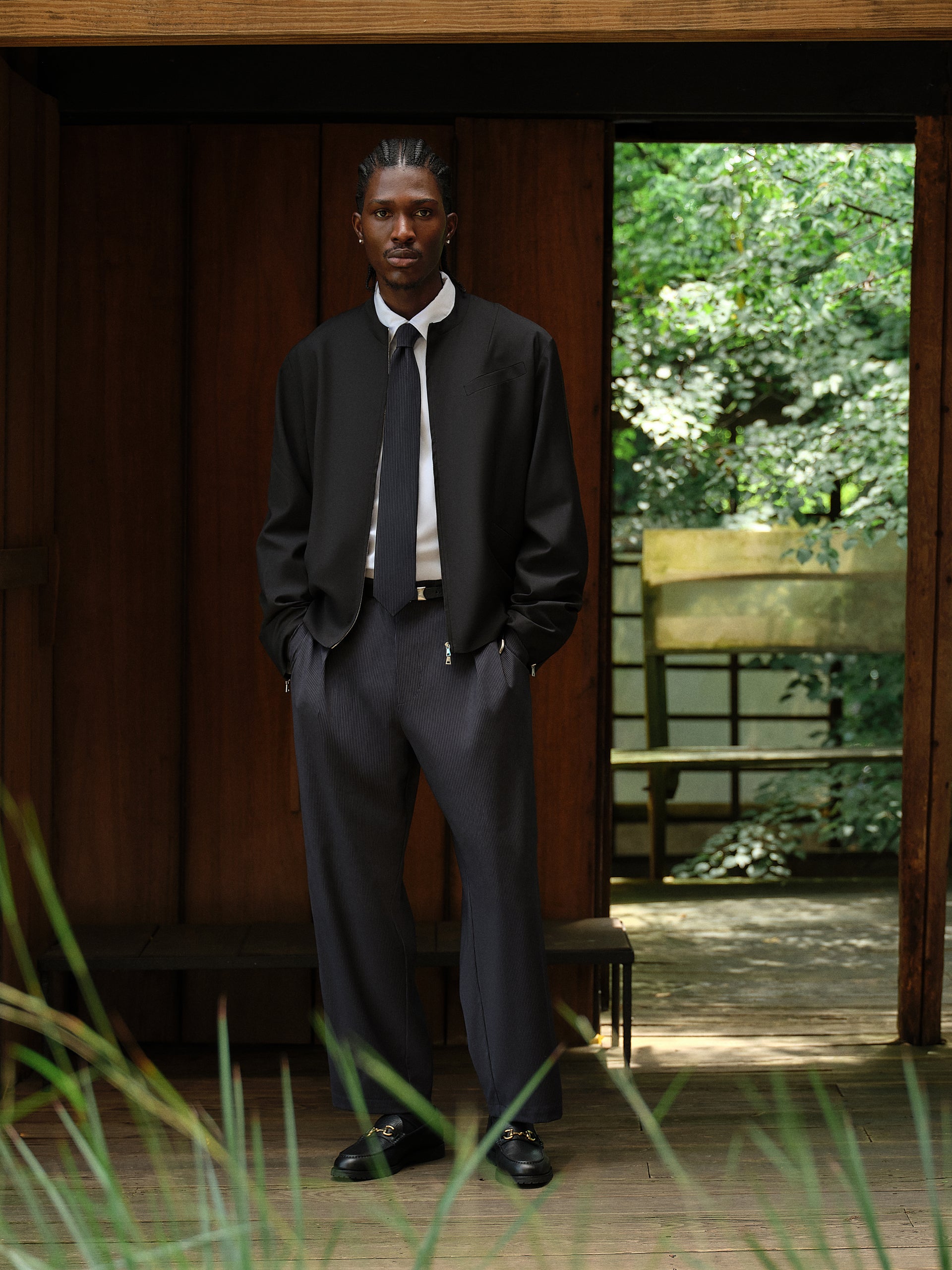 Model in a wood doorway wearing a black zip bomber, white shirt and tie, navy pinstripe trousers and black horsebit loafers