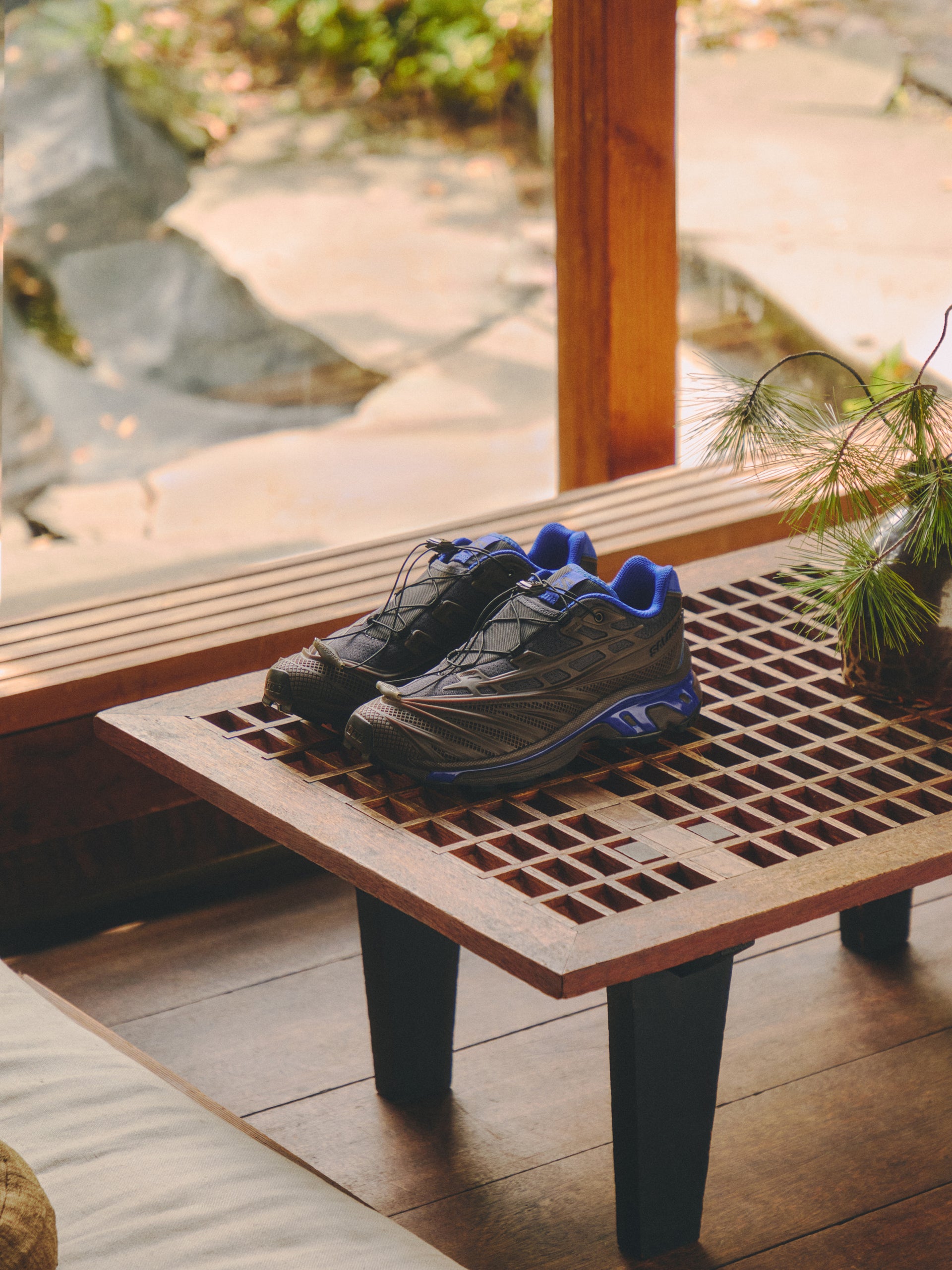 Salomon XT-6 sneakers in gray and cobalt blue on a wooden slat bench by a window overlooking a rock garden