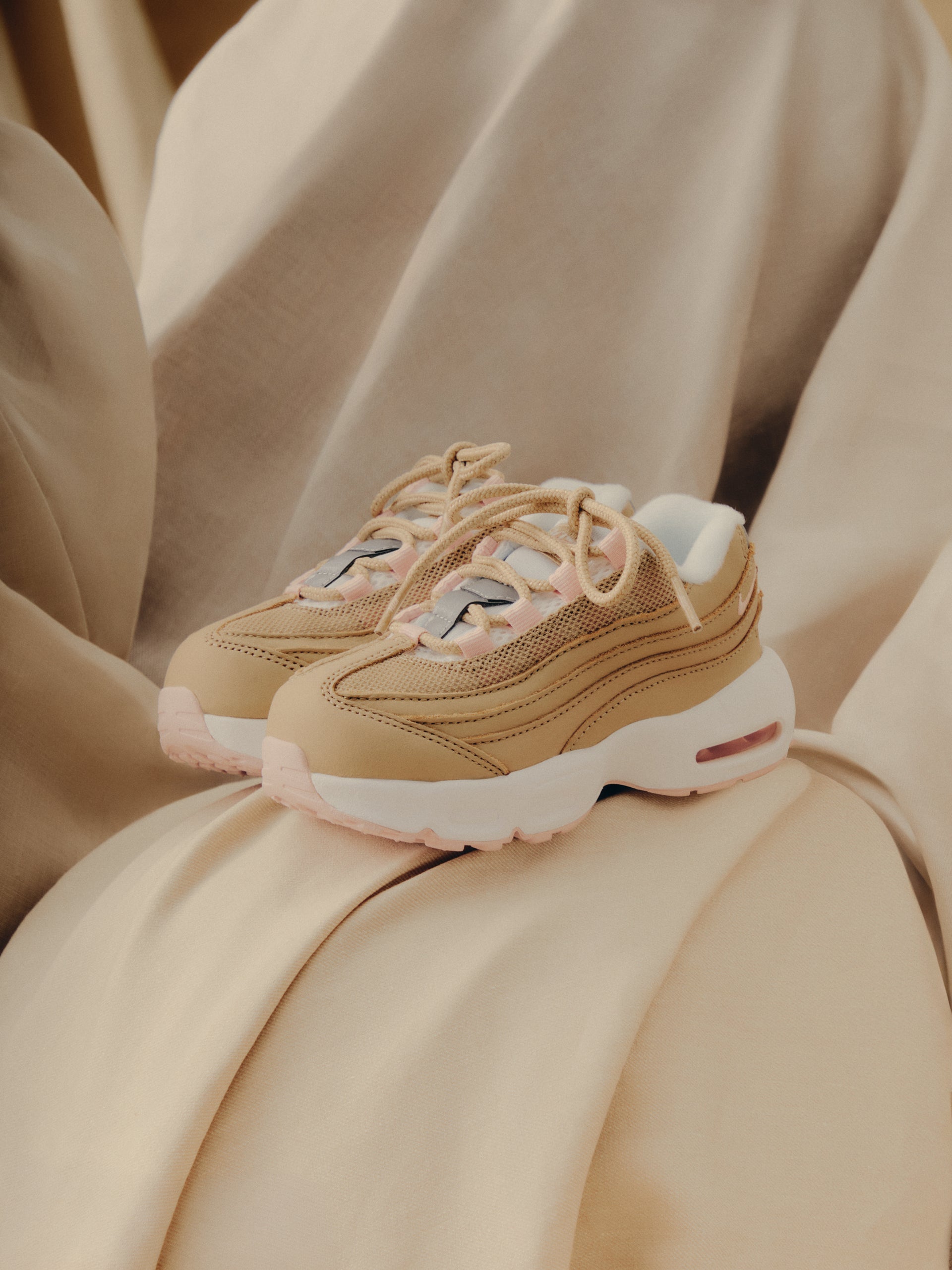 Pair of Nike Air Max 95 in tan with pink accents resting on soft beige fabric, Kith