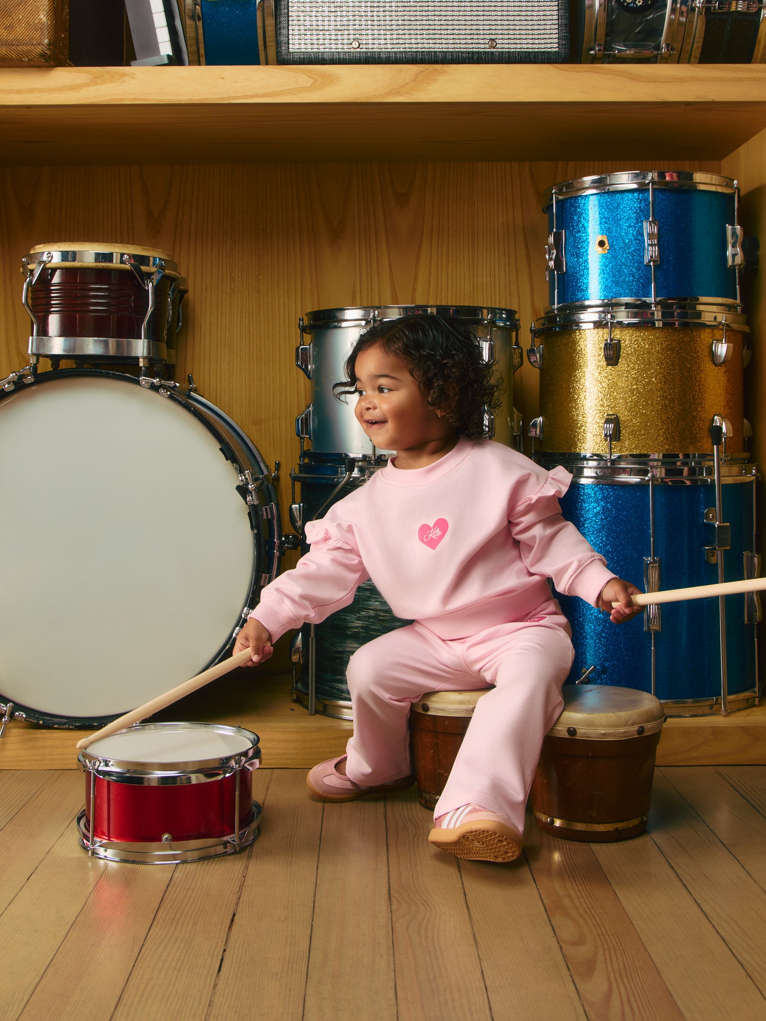 Toddler girl in pink Kith ruffle sweatshirt with heart logo and pink pants drums a red toy snare, glittery drum kit behind.