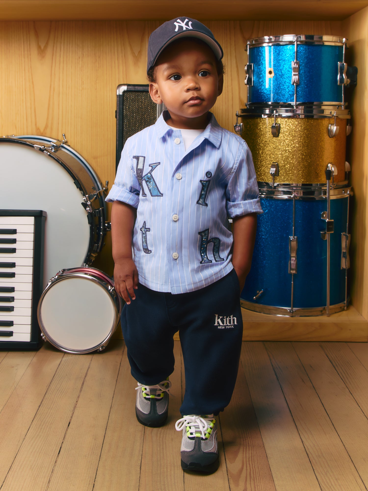 Toddler boy in navy Yankees cap, blue pinstripe kith appliqué shirt and navy Kith New York sweatpants by drums and piano.