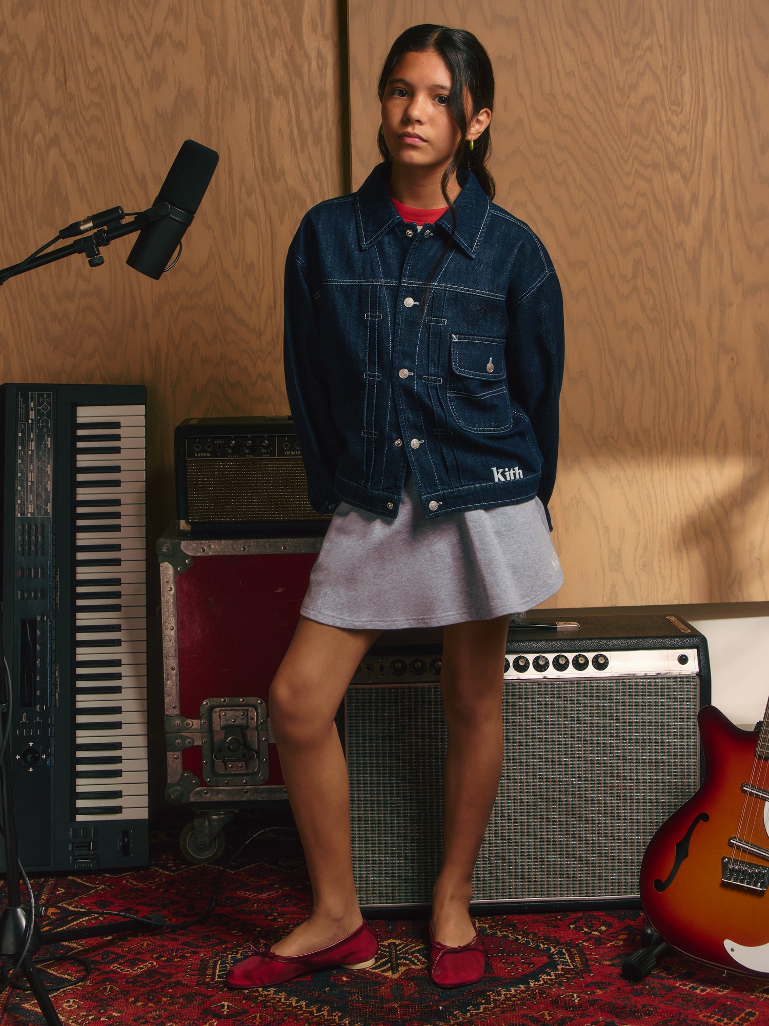 Girl in oversized indigo Kith denim jacket over gray skirt and red ballet flats beside a keyboard and amps in a studio.