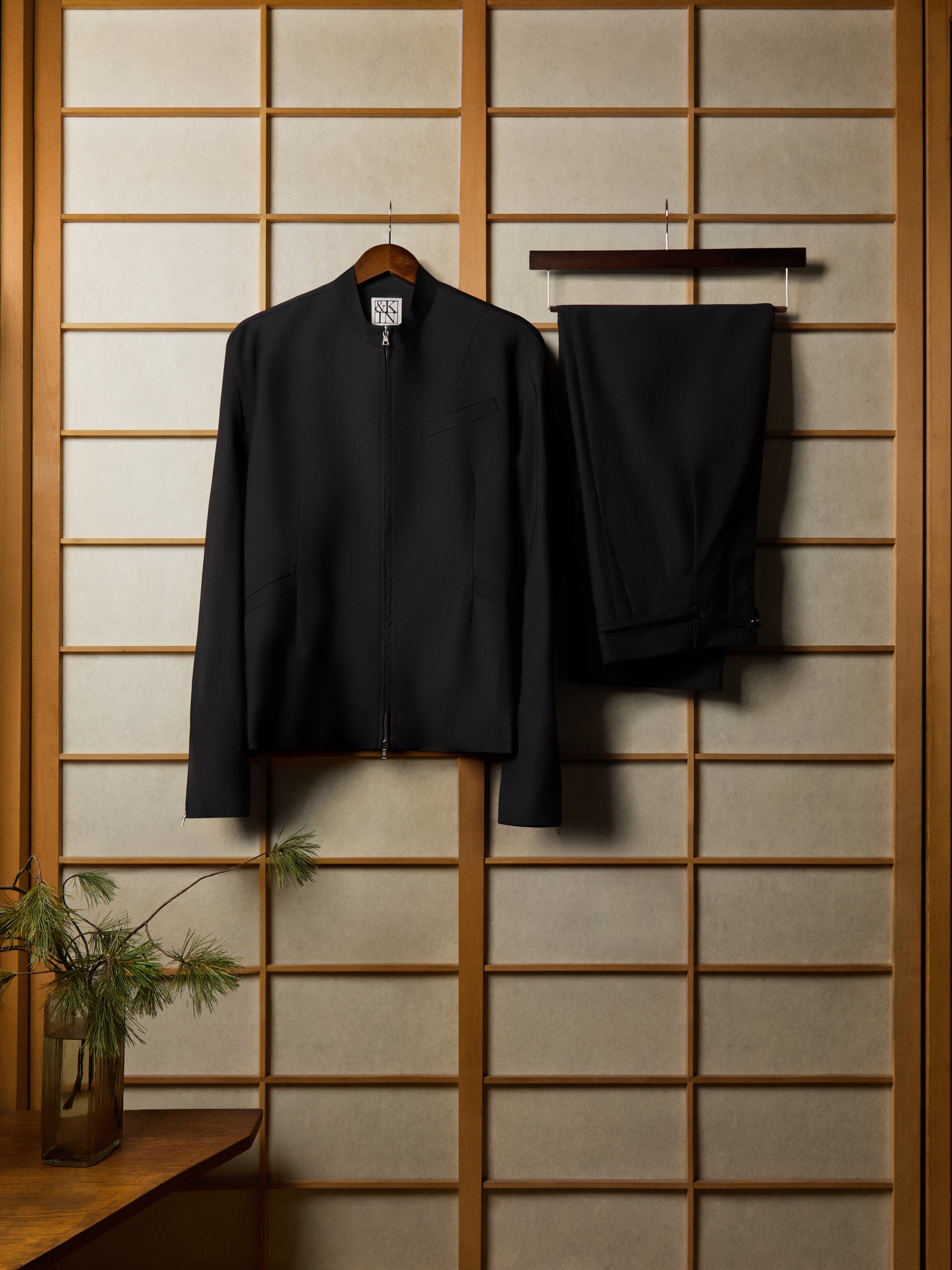 Black zip-up jacket and matching tailored trousers hung on a wooden shoji screen with pine sprig