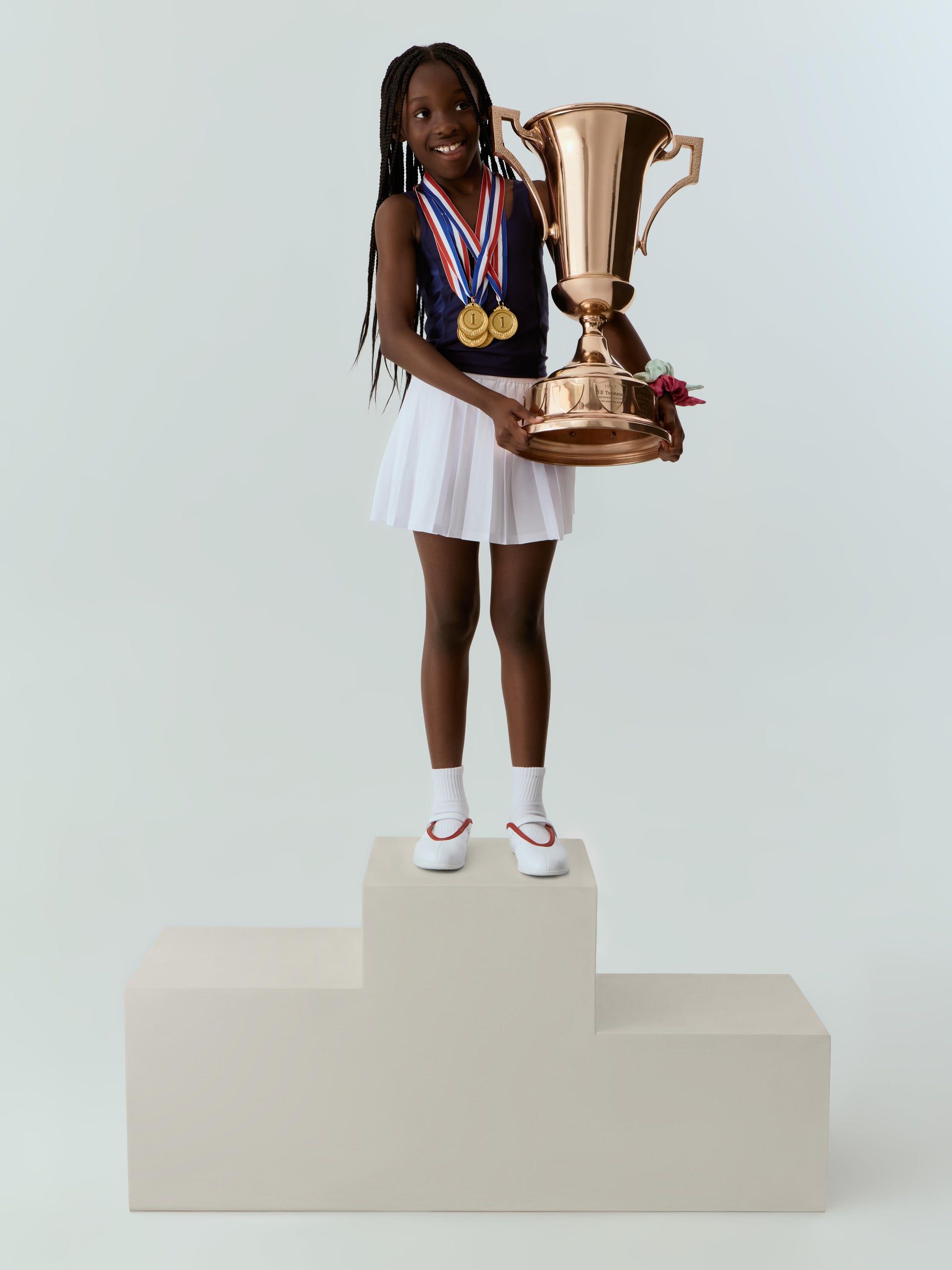 Girl in navy Kith New York tank and white tennis skirt resting a hand on a bronze trophy on a podium, medals in hand.