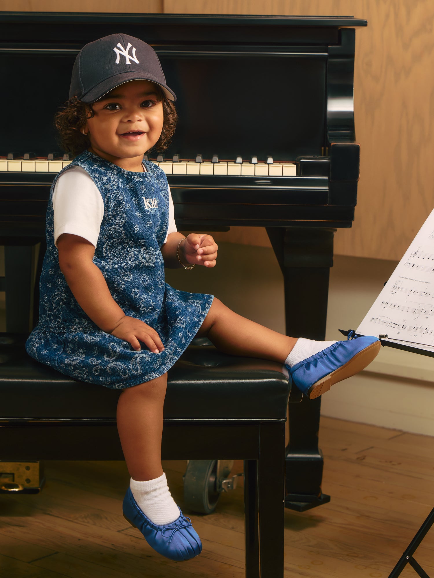 Smiling toddler in navy Yankees cap and blue paisley denim Kith dress with blue satin flats seated on a piano bench.