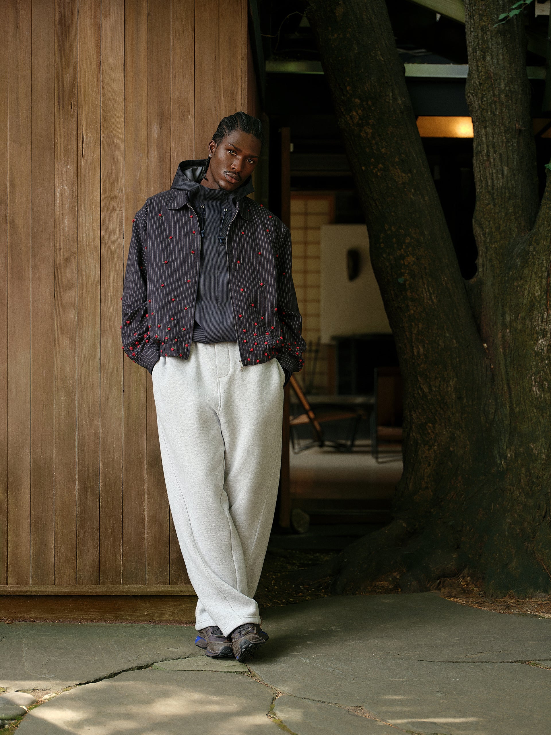 Model in navy pinstripe rose-embroidered jacket, ribbed hoodie and cream sweatpants against wood siding
