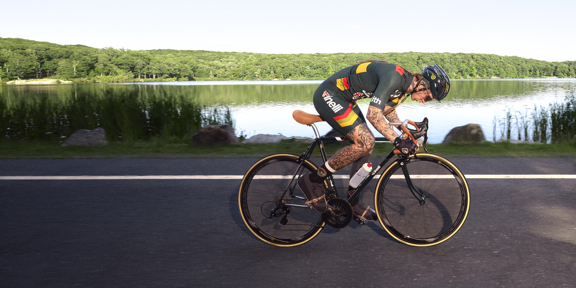 Cyclist in green Kith x Cinelli kit riding a road bike along a lake, side profile in motion