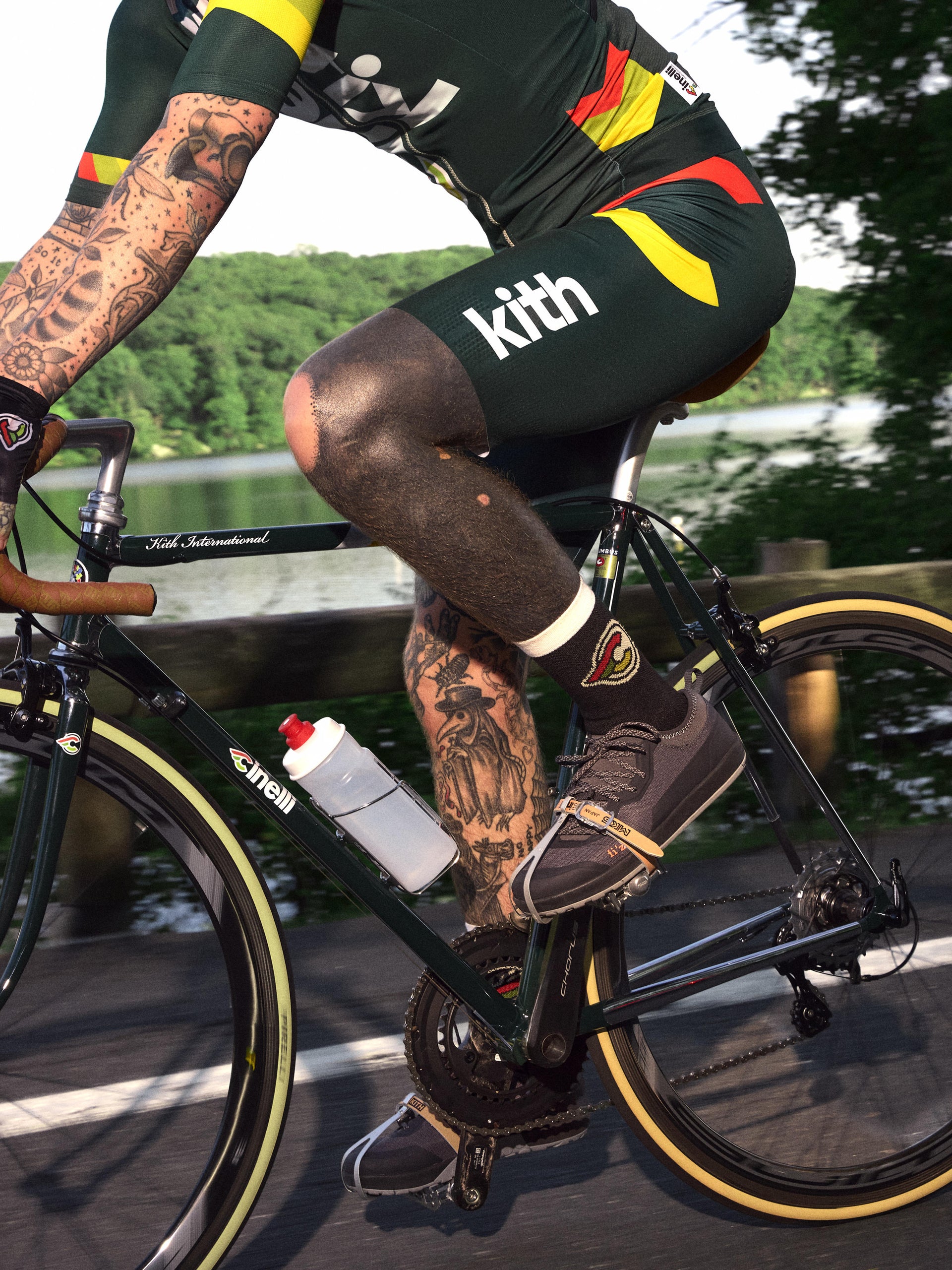 Detail of cyclist pedaling a green Cinelli road bike in Kith bib shorts, tattooed legs in motion