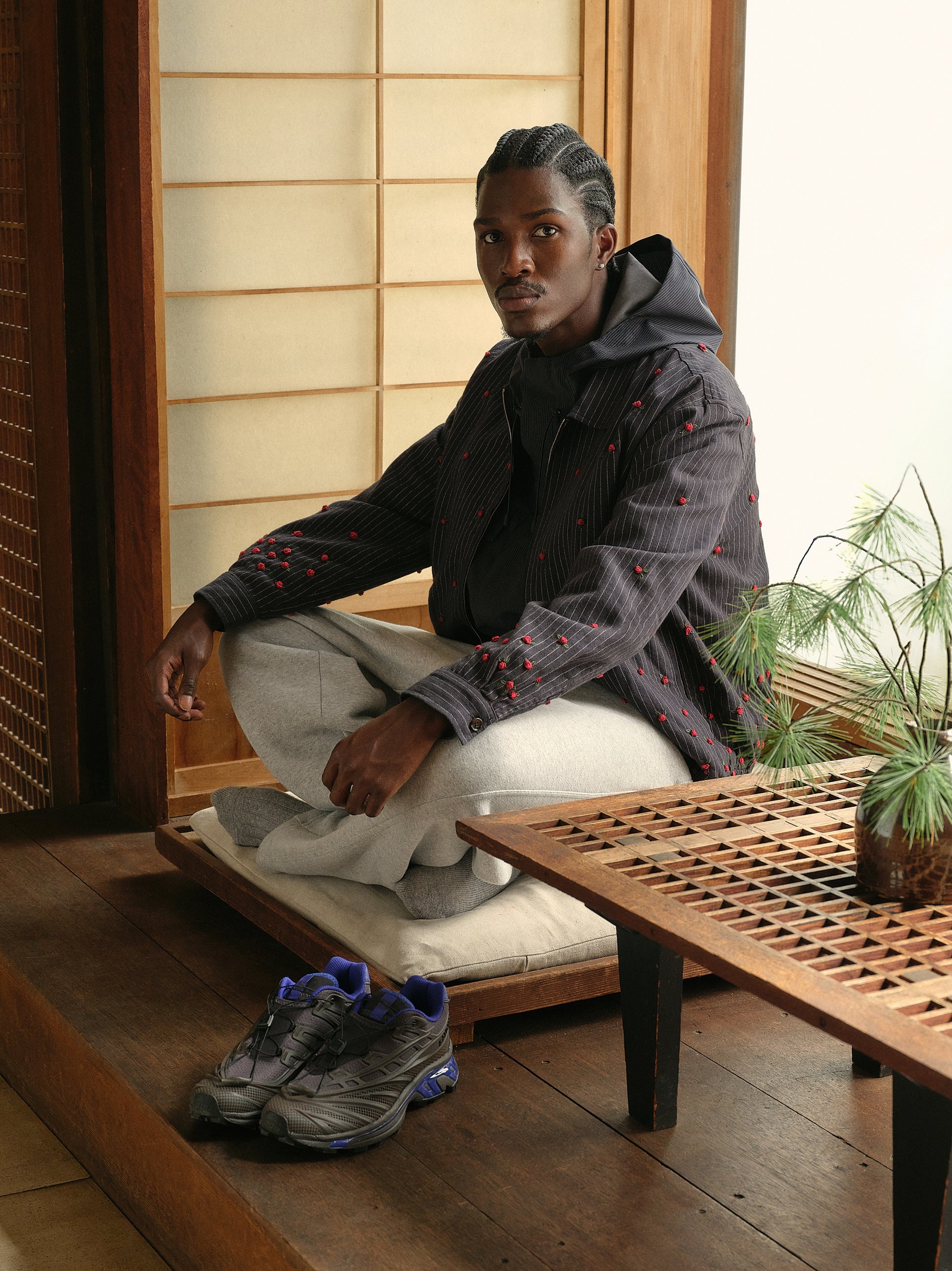 Model seated cross-legged in navy rose-embroidered pinstripe jacket and gray sweatpants, Salomon sneakers nearby