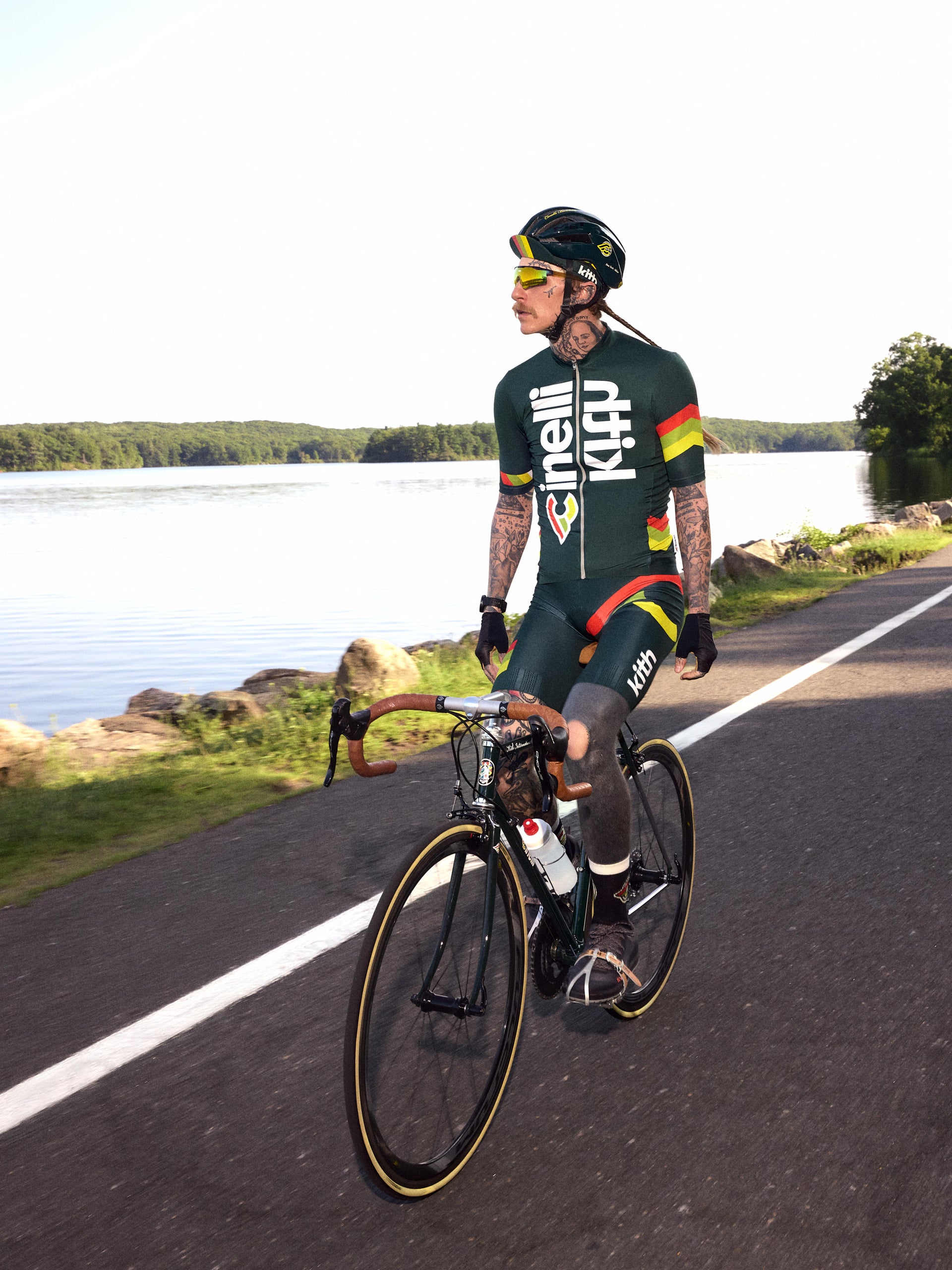 Tattooed cyclist riding a green Cinelli road bike lakeside in Kith x Cinelli green cycling kit and helmet, front view