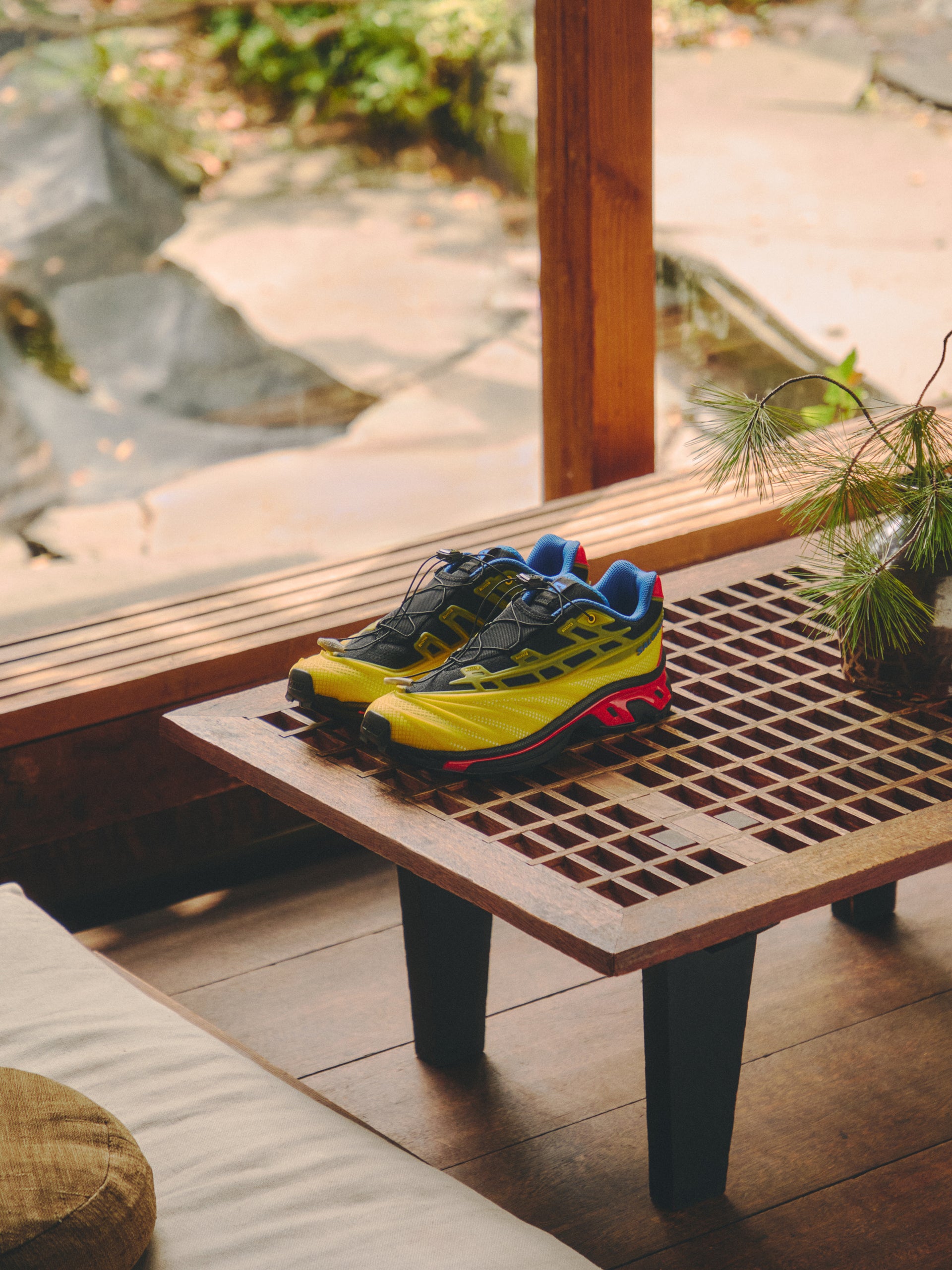 Salomon XT-6 sneakers in yellow, black, blue and red on a wood slat bench, Japanese garden and pine sprig behind