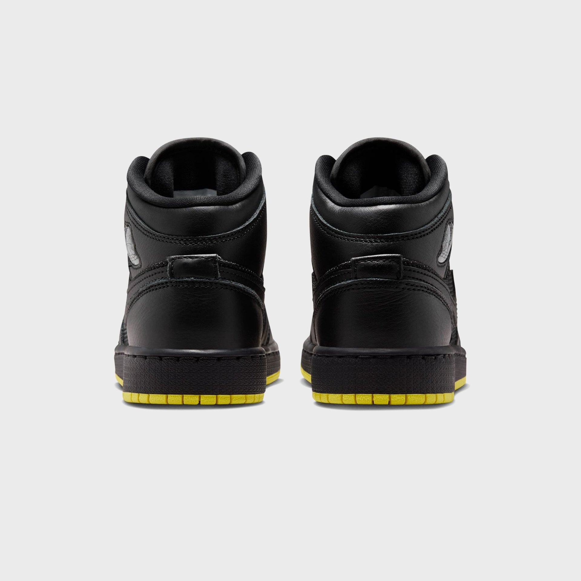 Air Jordan 1 Mid SE pair, rear view of black leather heels with silver Wings logos above opti yellow outsoles