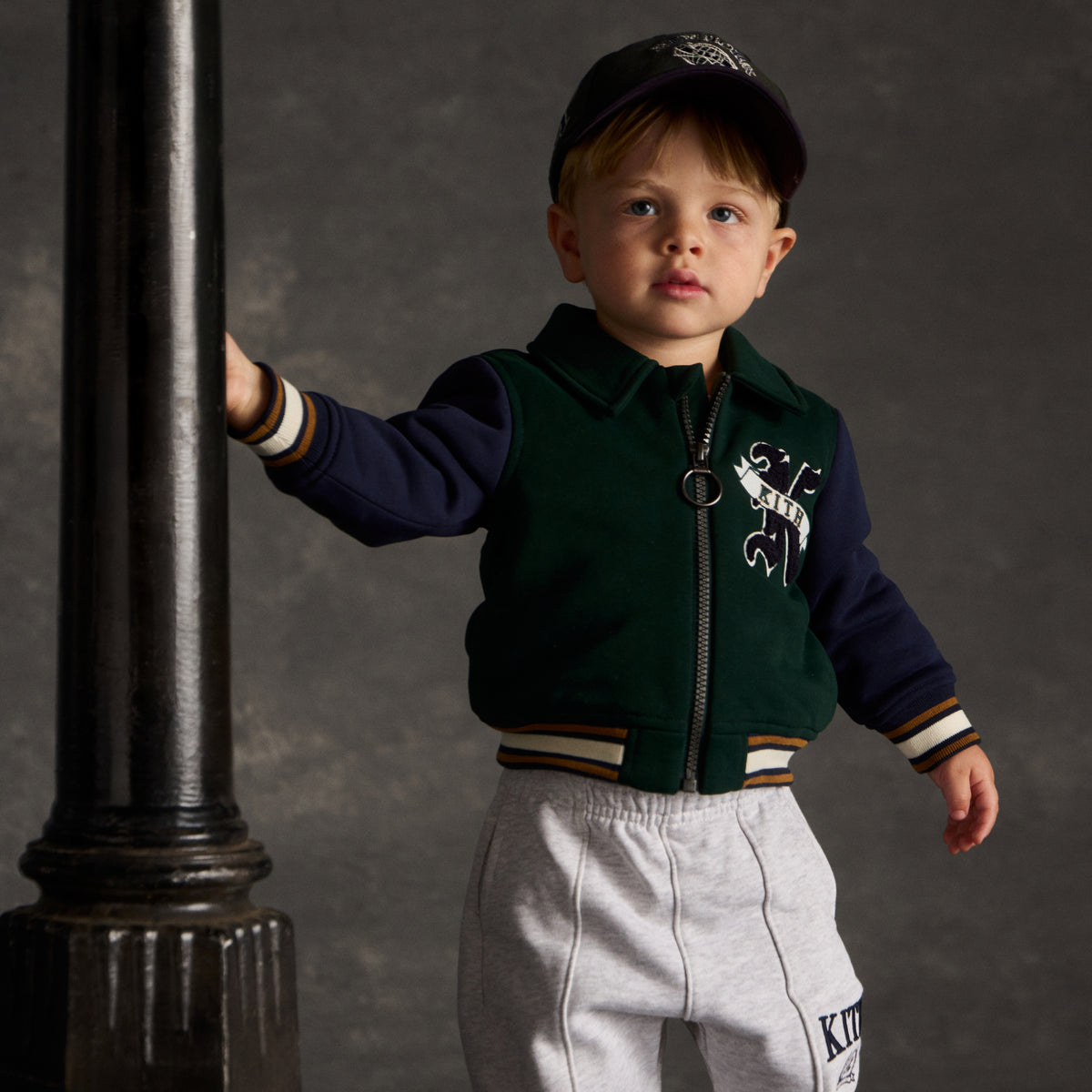 Toddler boy in a navy New York cap and green colorblock Kith Kids zip jacket with K logo patch, holding onto a lamppost