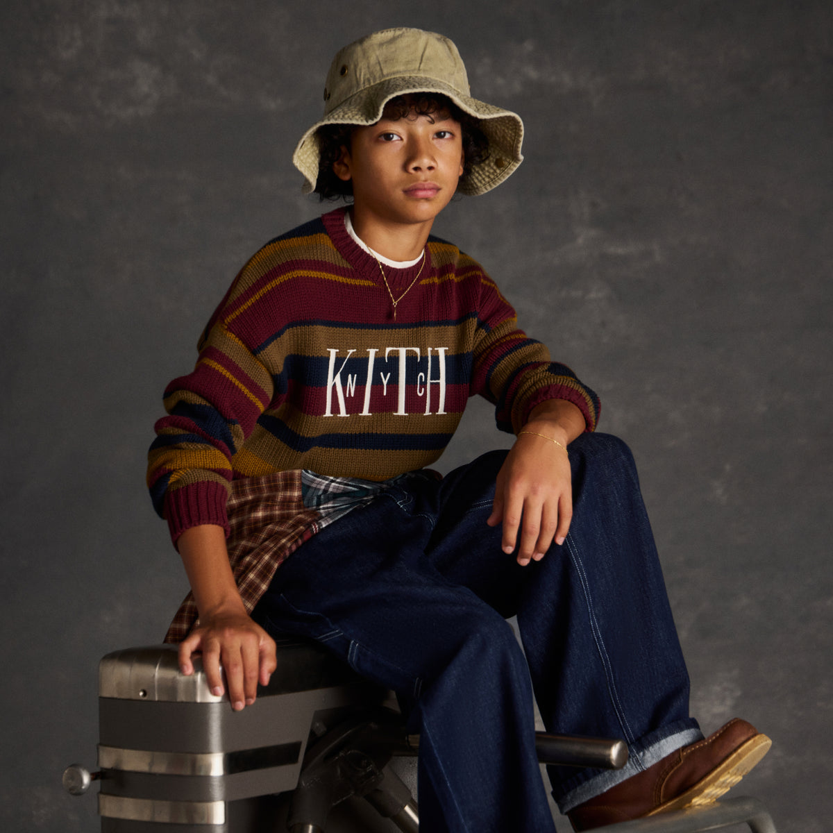Boy in a khaki bucket hat and striped burgundy, navy, and mustard Kith NYC sweater, seated on metal luggage cases