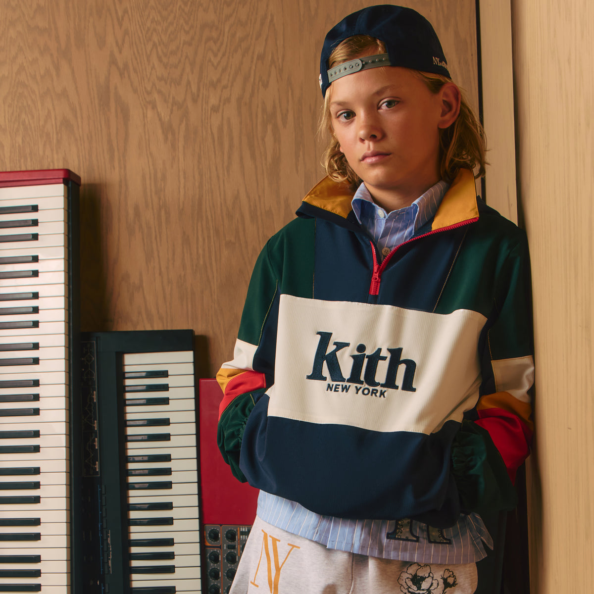 Boy in a navy New York cap and green, navy, and cream colorblock Kith New York quarter-zip jacket, standing beside upright keyboards