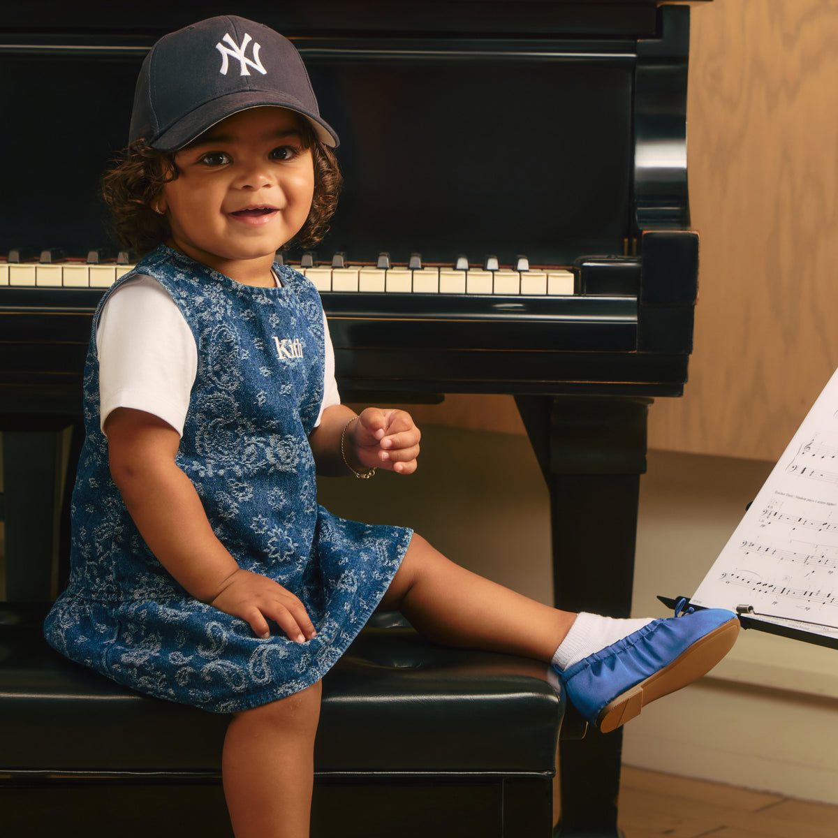 Toddler girl in a navy New York Yankees cap and blue paisley denim Kith jumper dress, seated at a piano bench