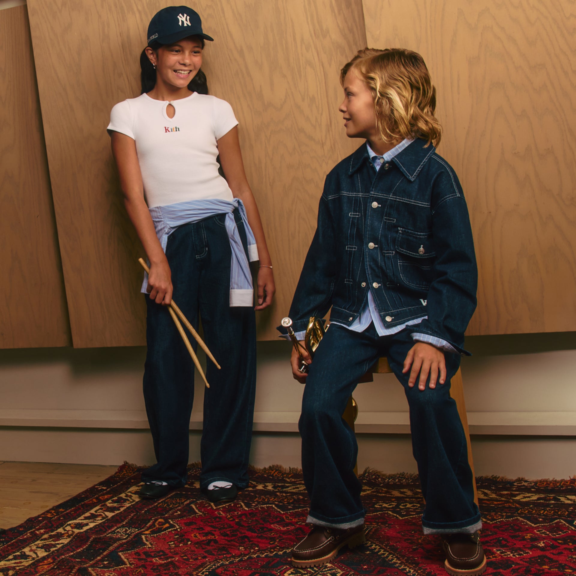 Two kids by wood paneling: girl in a navy Kith Yankees cap and white logo tee holding drumsticks, boy in an indigo denim jacket with a trophy