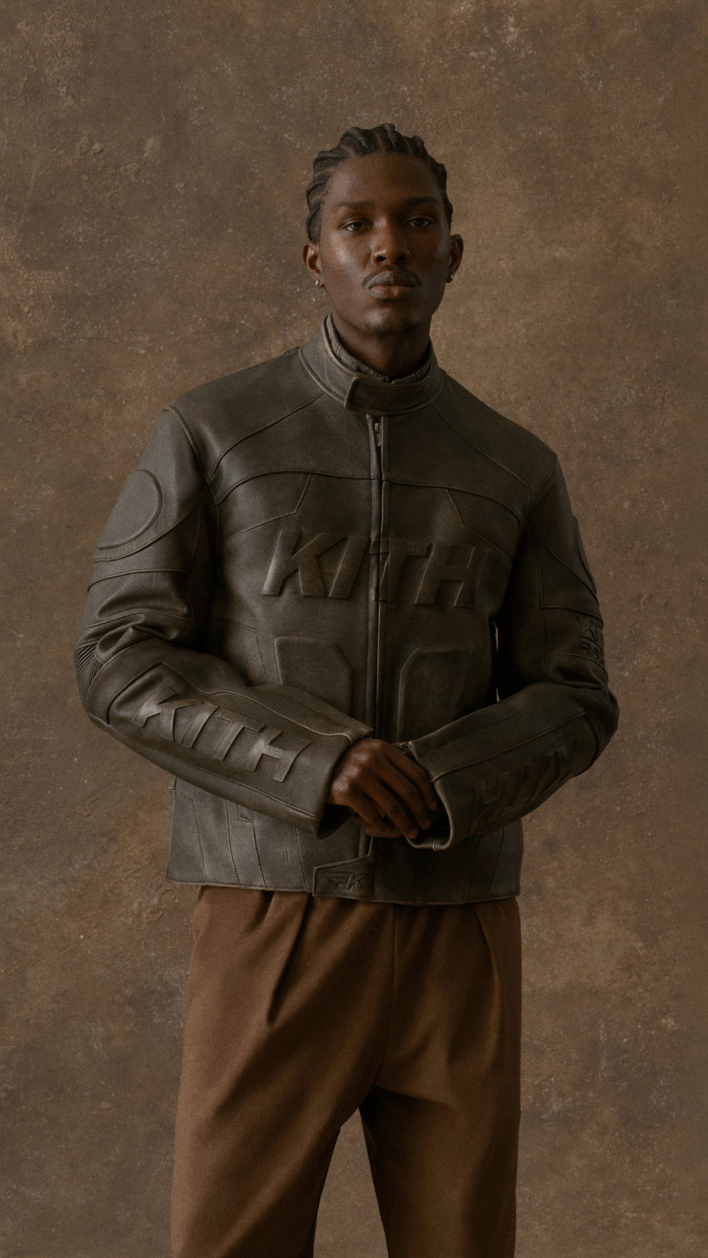 Full-body view of a man in a brown leather Kith jacket and brown trousers standing against a brown textured backdrop