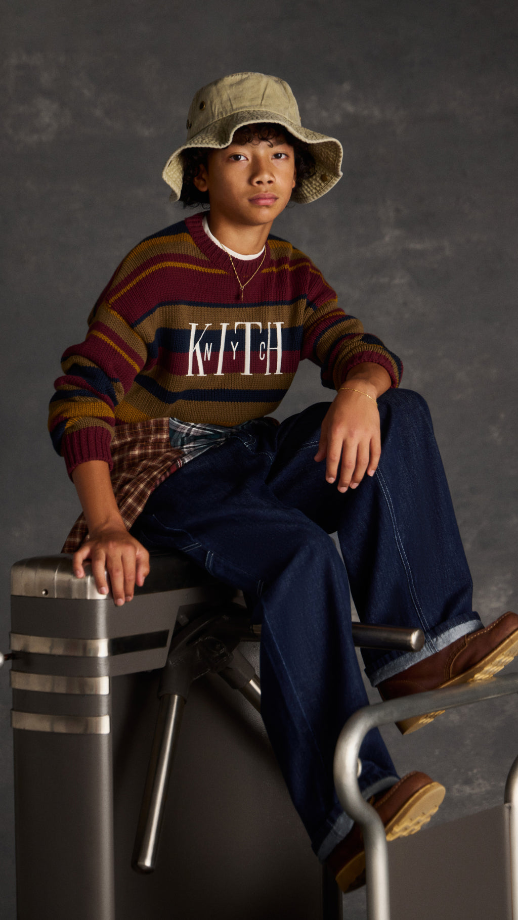 Boy in a khaki bucket hat and striped burgundy, navy, and mustard Kith NYC sweater, seated on a subway turnstile in dark jeans