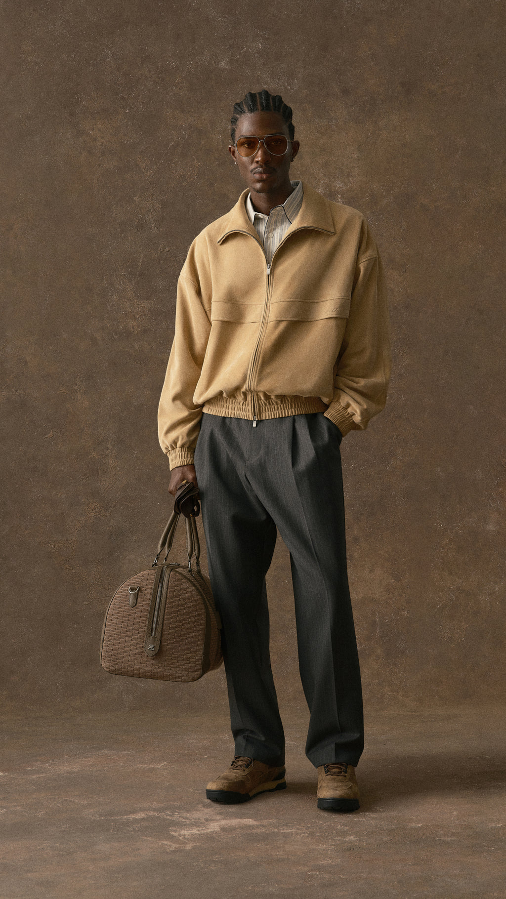 Mobile hero image of a man in a tan Kith zip jacket and dark trousers holding a brown leather bag against a brown backdrop