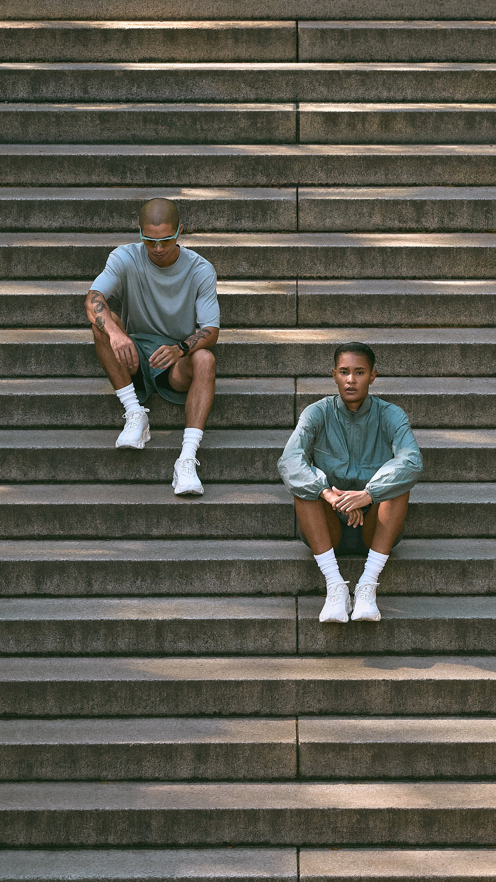 Two men wearing light blue-gray Kith athletic wear seated on concrete stadium steps, mobile crop