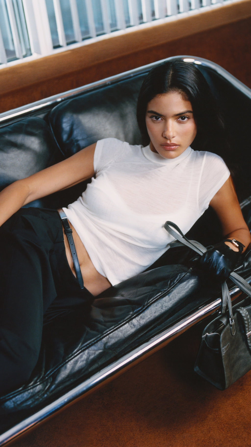 Model reclining on a black leather bench seat wearing a white t-shirt and black trousers, holding a black leather bag, mobile crop