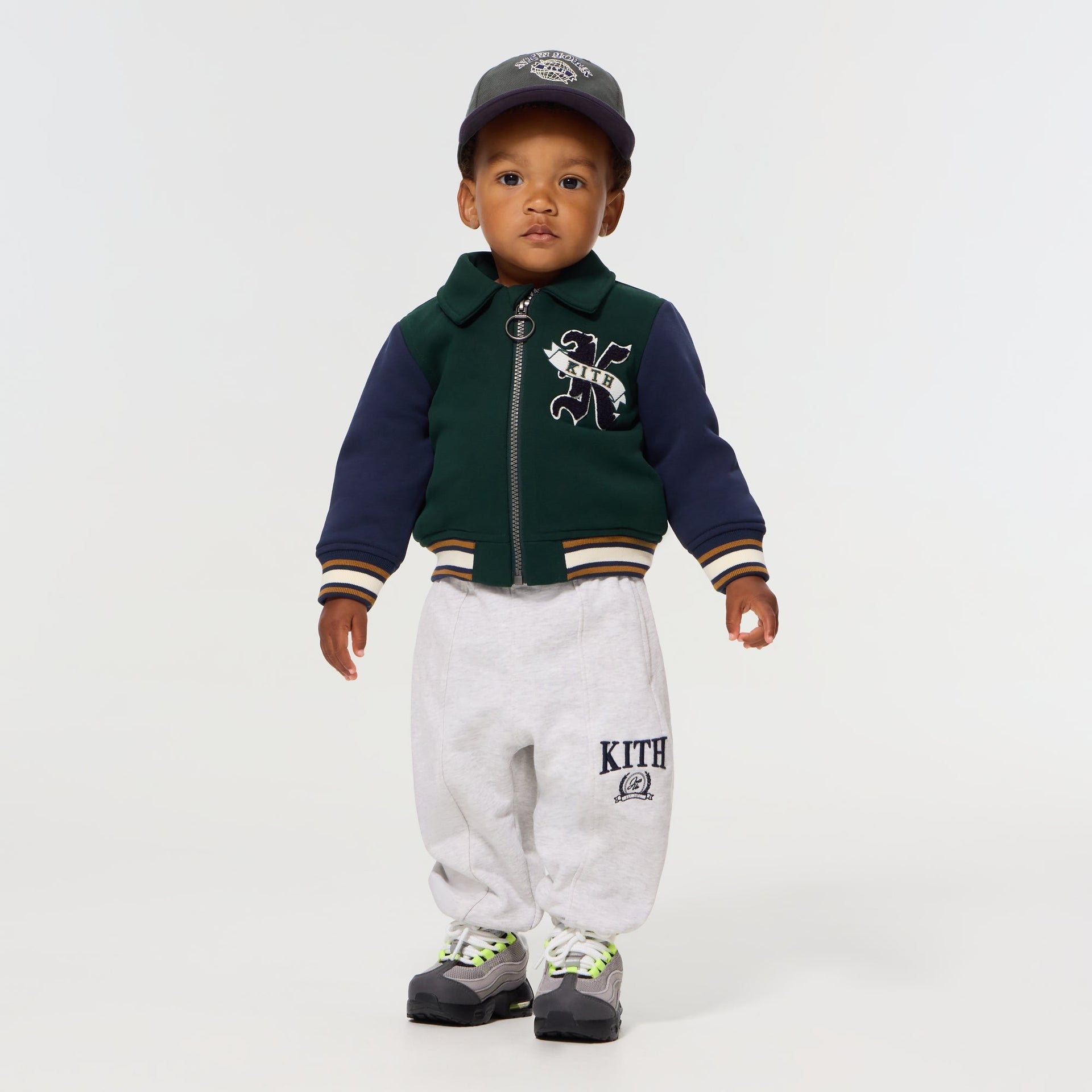 Baby wearing the Kith Baby Varsity Jacket in Stadium with a New York cap, grey Kith sweatpants and sneakers