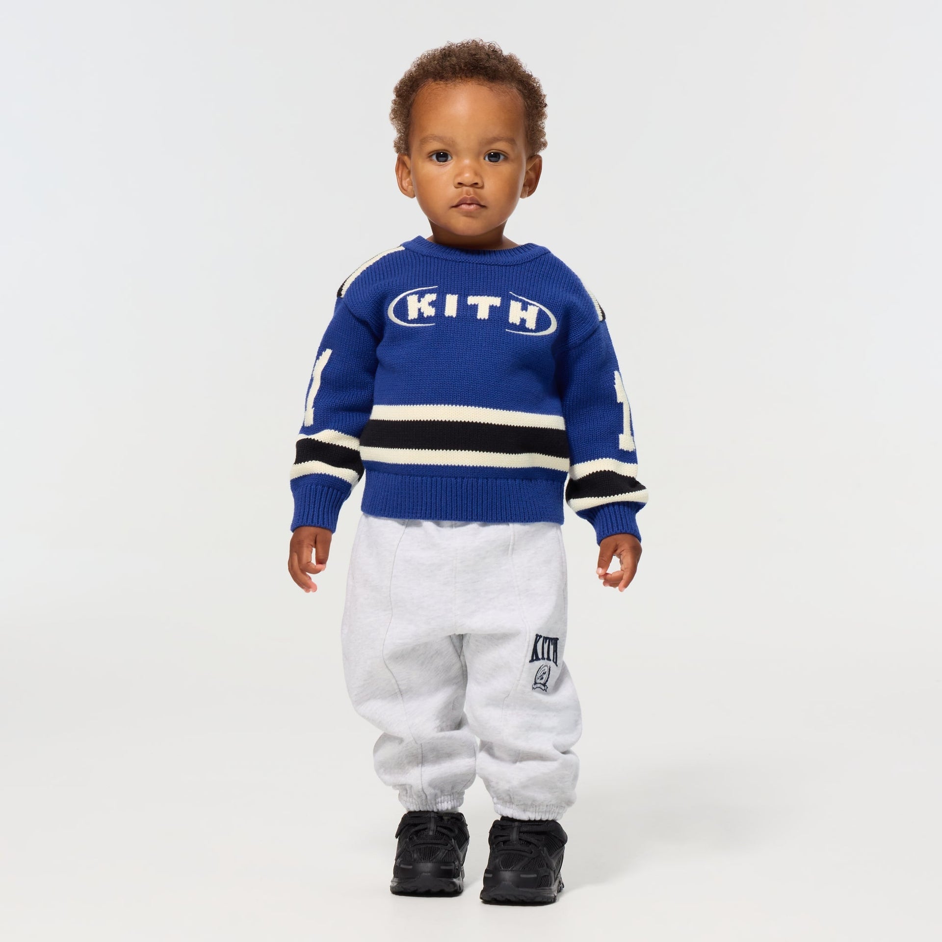 Toddler wearing the Kith Baby Varsity Nelson Knit Crewneck in Cyanotype with heather grey Kith sweatpants and black sneakers