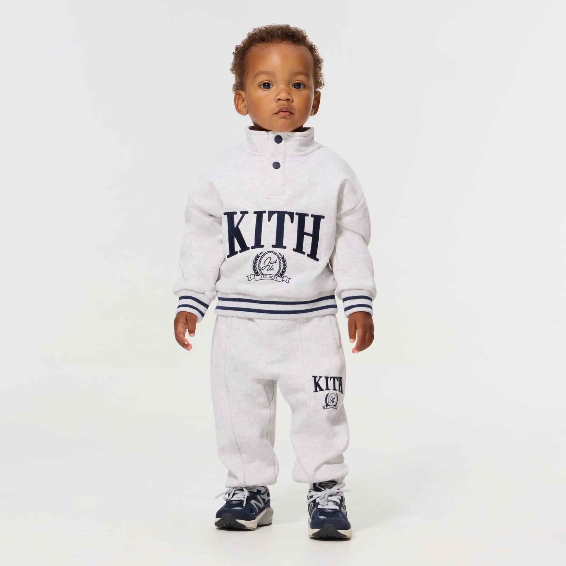 Baby wearing the Kith Baby Collegiate Snap Hunter Crewneck in Light Heather Grey with matching sweatpants and navy sneakers
