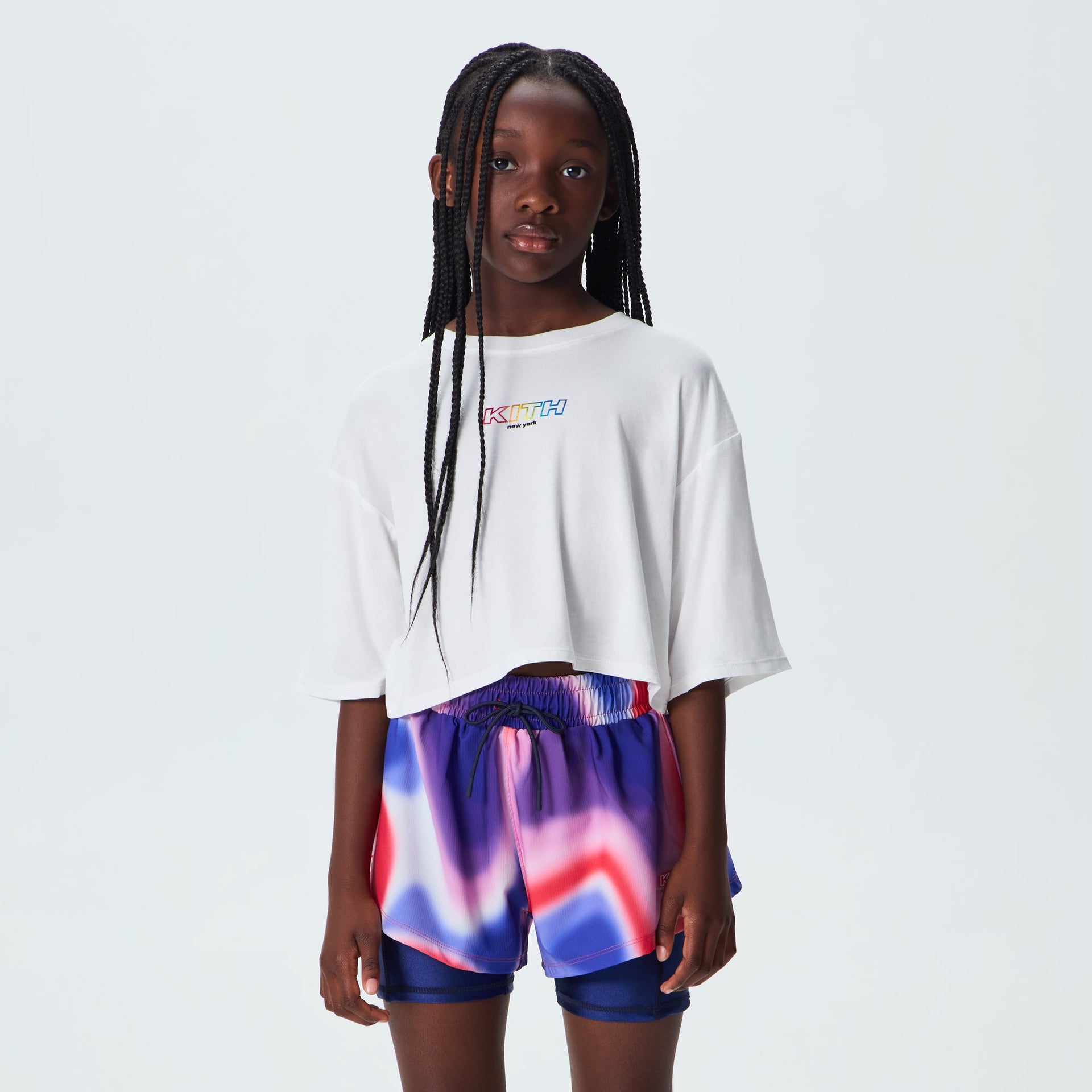 Girl wearing white Kith Kids Fei Cropped Tee with rainbow logo, styled with tie-dye shorts, on-model front view
