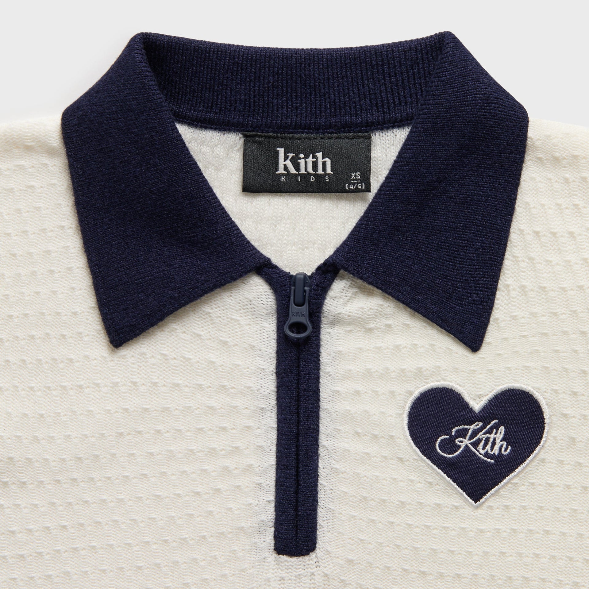 Close-up of navy collar, quarter-zip and navy heart Kith patch on Silk cream Kith Kids Taylen Crop Polo