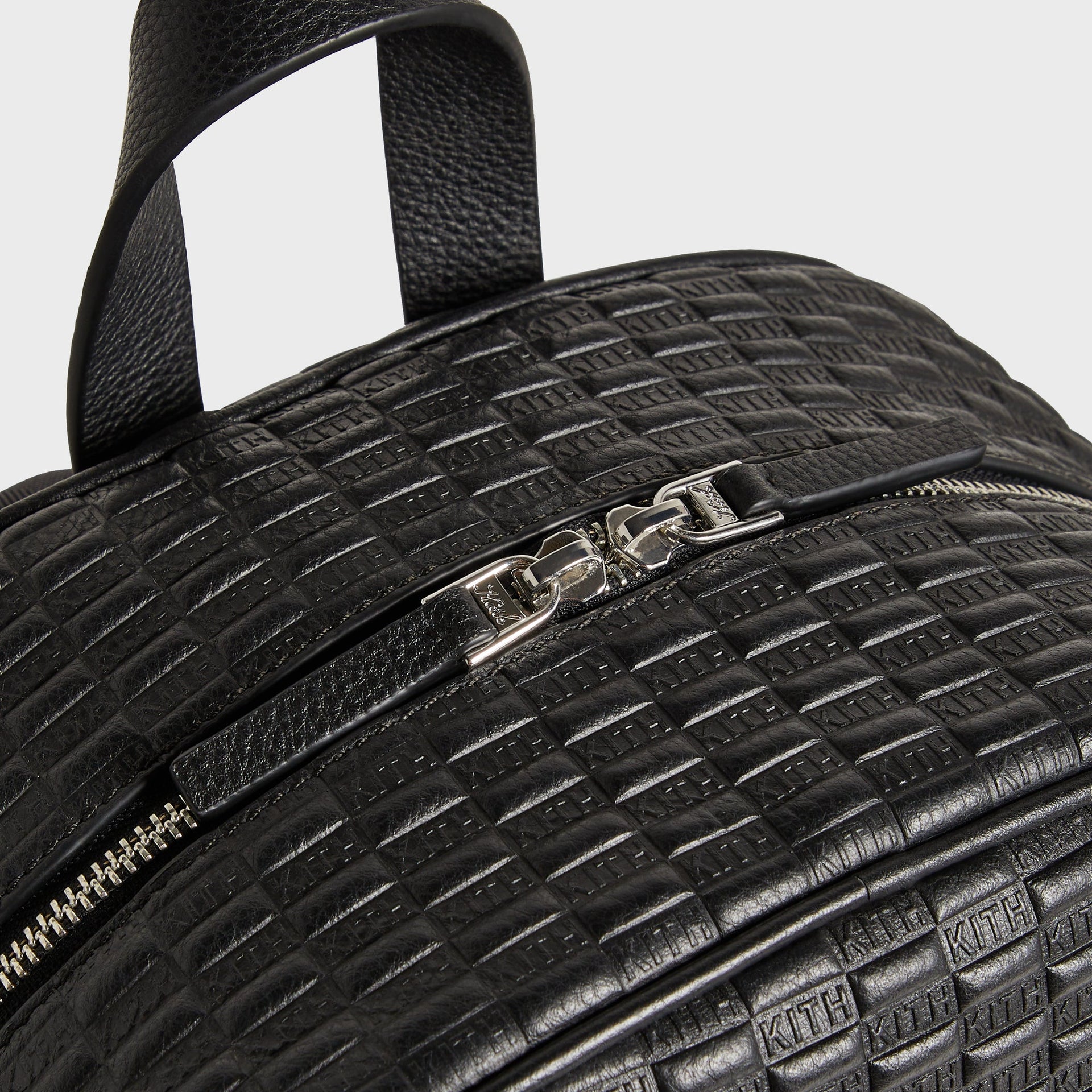 Close-up of the Kith Monogram Tumbled Leather Backpack's top handle, side zip pull, and adjustable strap buckle hardware.