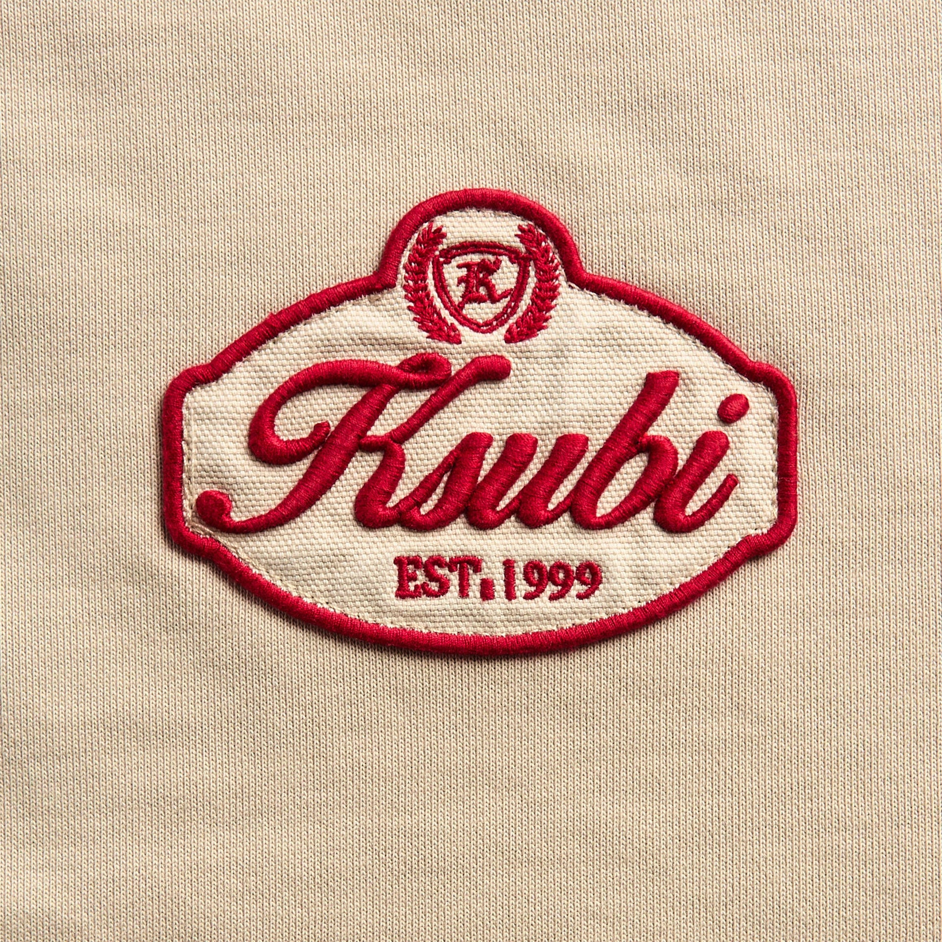 Close-up of the burgundy embroidered Ksubi EST. 1999 crest patch on the beige hoodie fabric