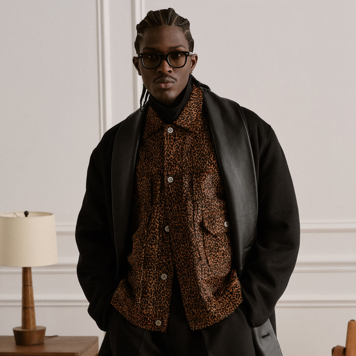 Man in black wool overcoat and leopard-print button-up shirt with black turtleneck, glasses, and braids, indoor portrait