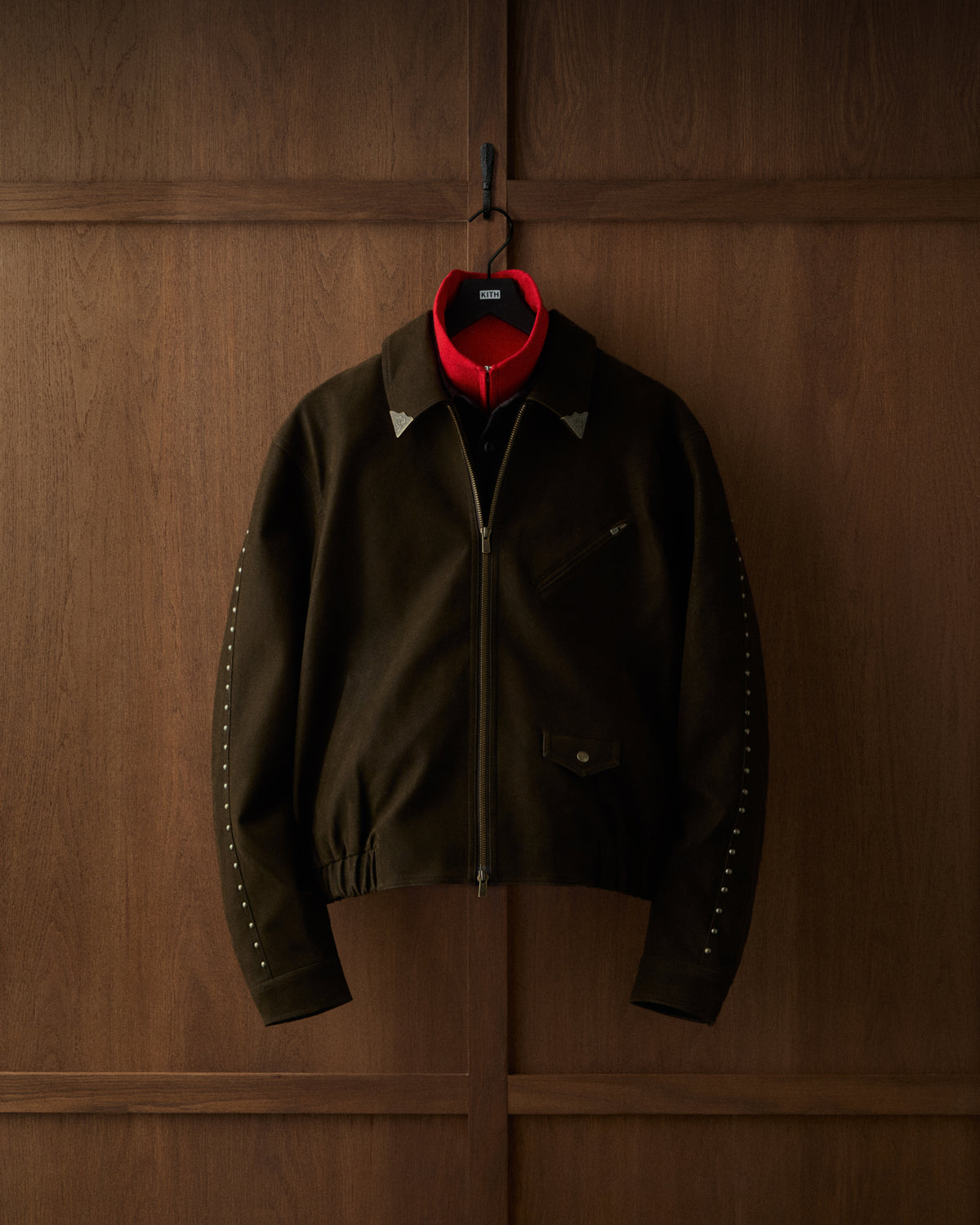 Close-up of a dark olive Kith jacket with silver stud details on the sleeve, hanging against dark wood paneling